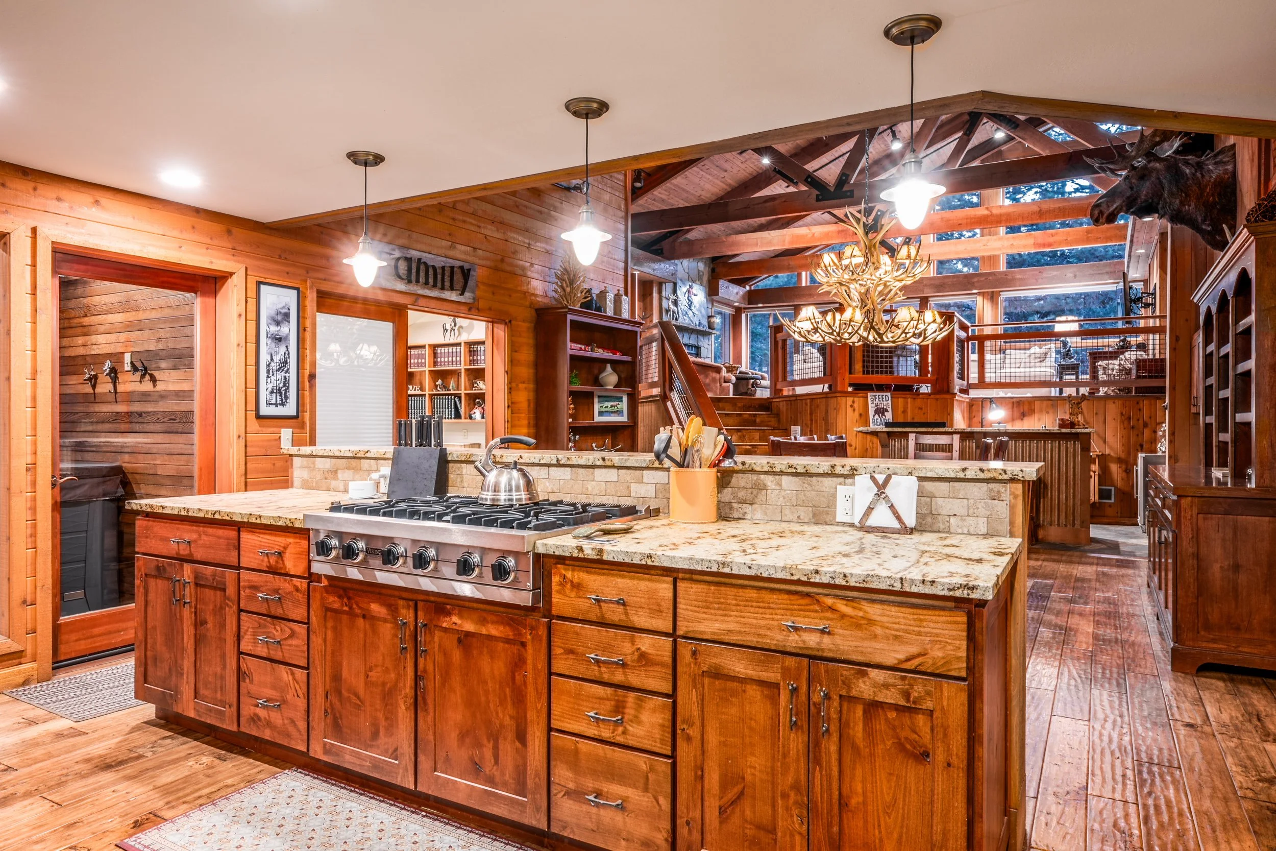 Interior view of a rustic kitchen with wooden cabinets, granite countertops, a stove, and decorative antler chandelier hanging from the ceiling. A mounted bear's head is visible on the upper right corner.