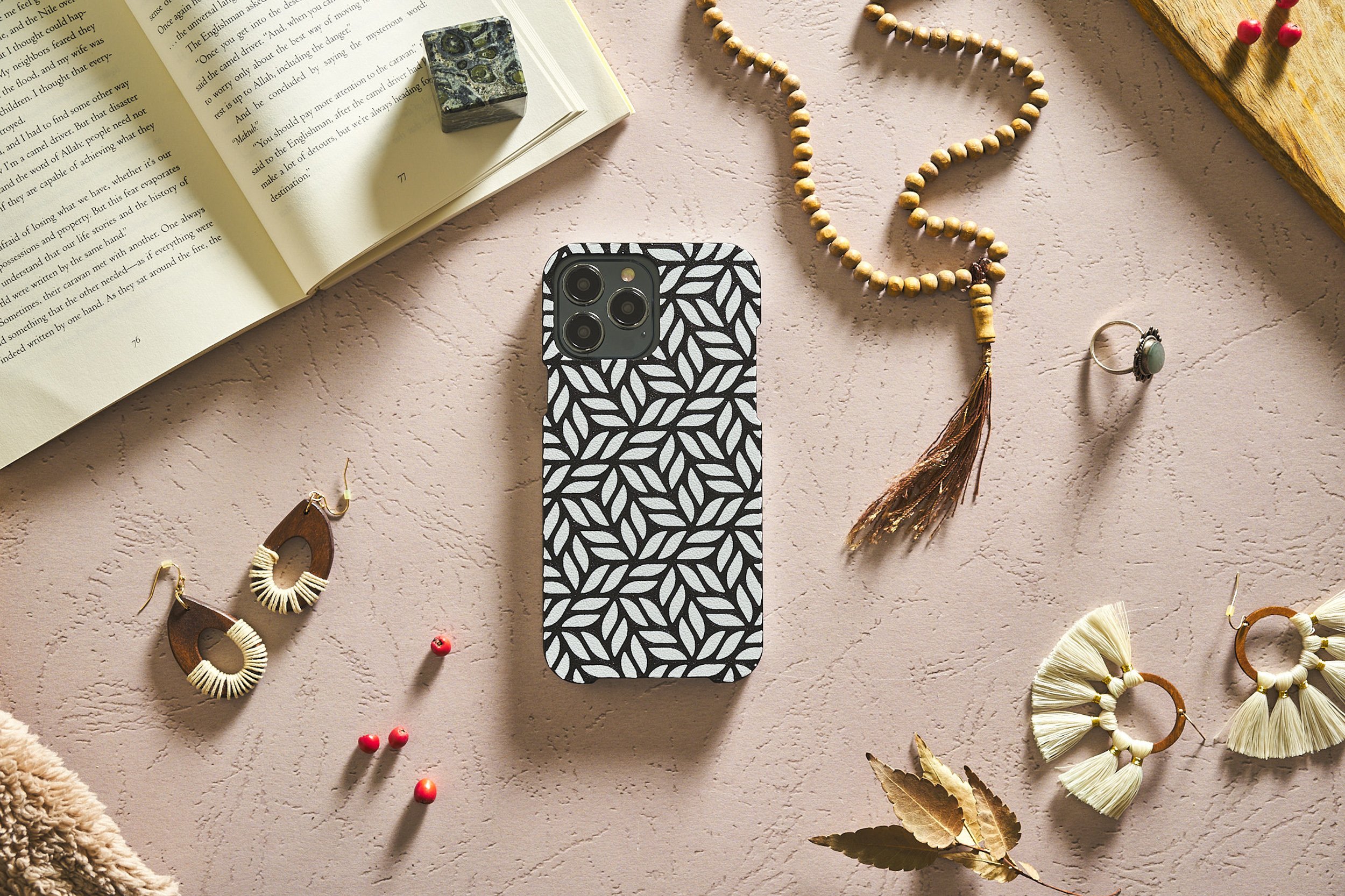 A flat lay of a smartphone with a black-and-white patterned case, an open book with text, various earrings, a beaded necklace, a ring, dried leaves, and red berry accents on a textured pink surface.