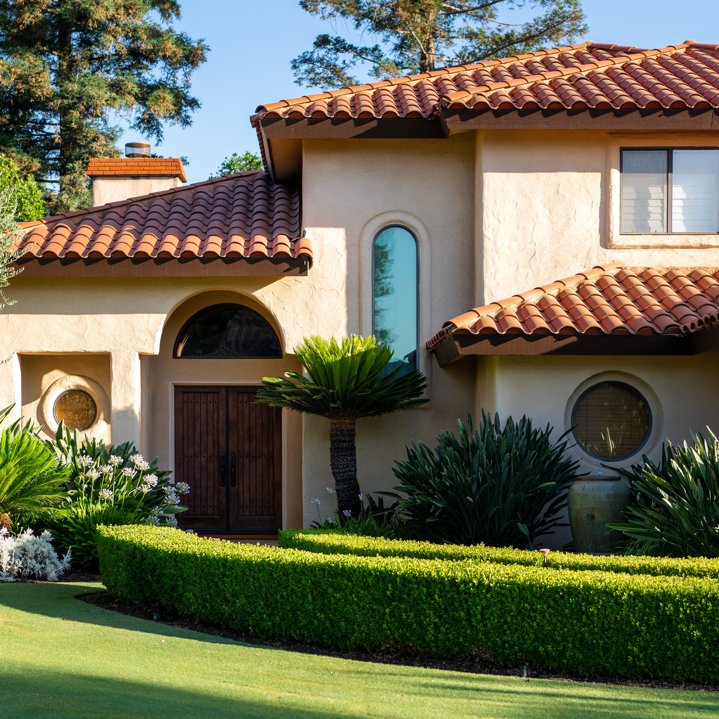 Mediterranean-style home with a red tile roof, arched wood entry, and manicured boxwood hedges in golden afternoon light