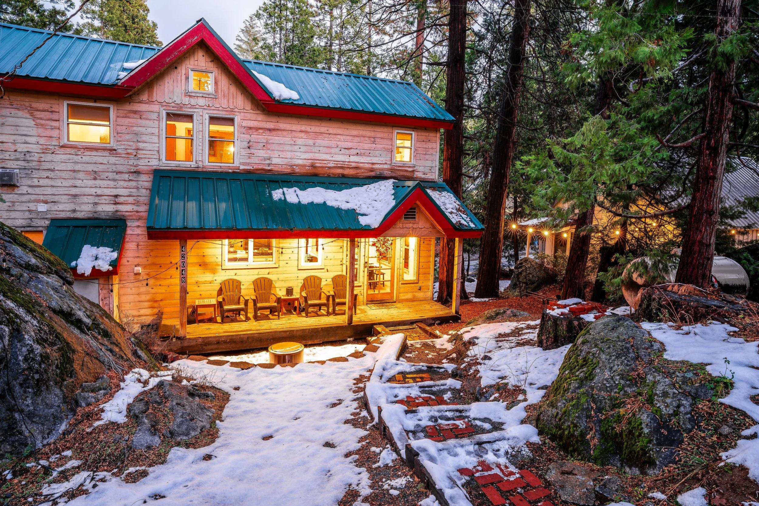 A cozy wooden house with snow on the roof is illuminated warmly from inside, situated among tall evergreen trees during dusk. The house features a small porch with four yellow chairs and string lights hanging nearby, with snow-covered rocks and a pat