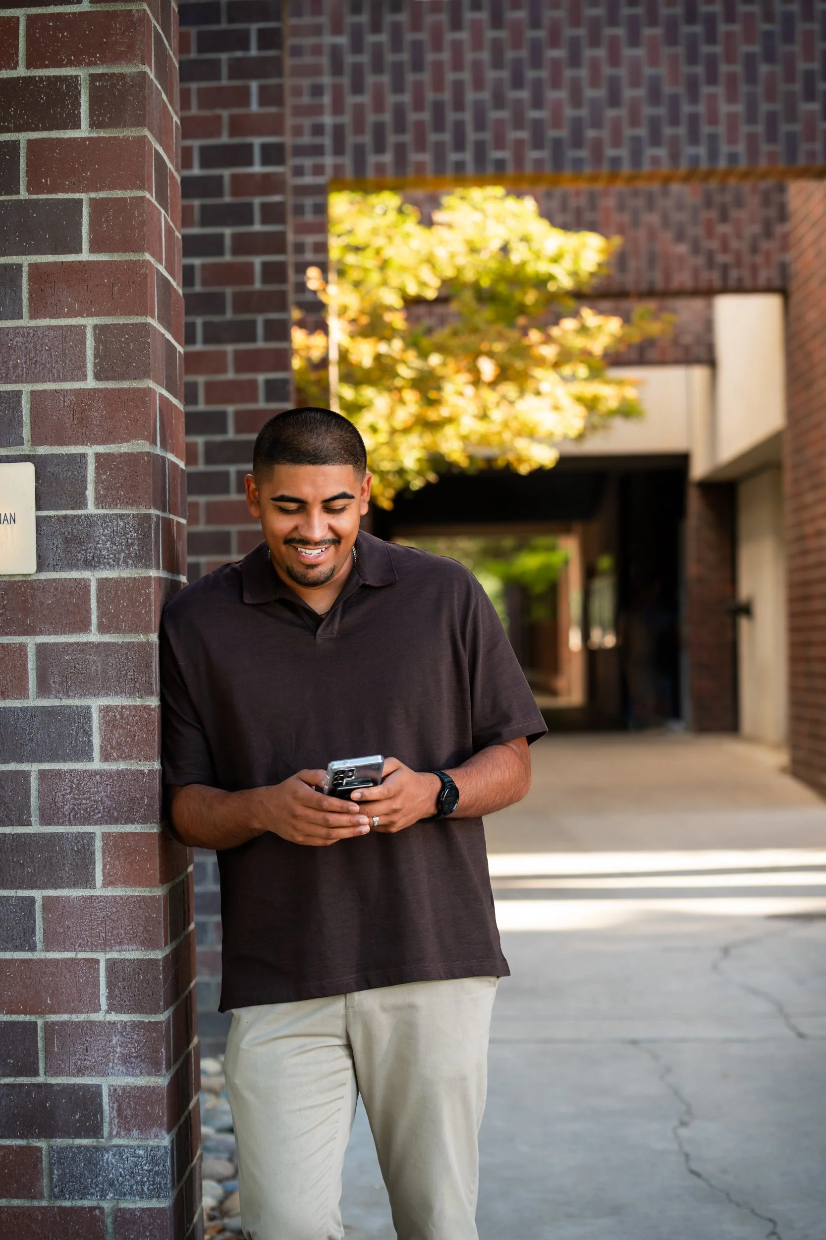 Real estate agent in a brown polo checking his phone outside a brick office building in warm afternoon light