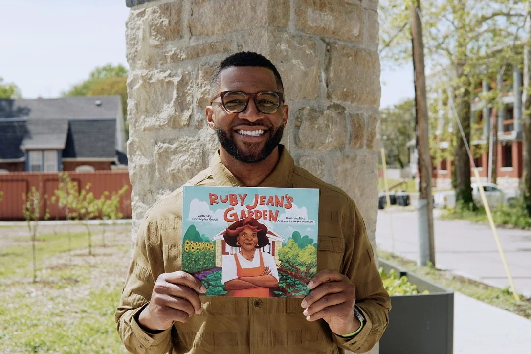 Chris Goode smiling and holding a children's book titled "Ruby Jean's Garden" in front of a stone wall outside.