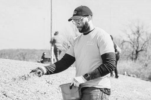 Chris Goode wearing a cap, glasses, and gloves stands outdoors, holding a gardening tool.