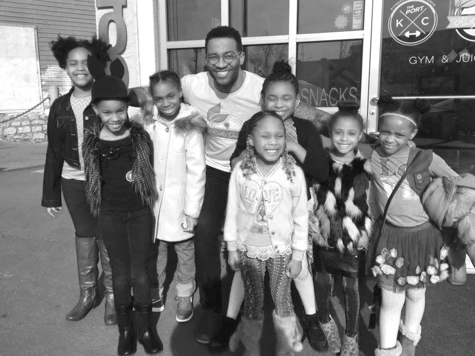 A group of nine children with Chris Goode smiling outdoors, standing in front of a gym and juice bar.