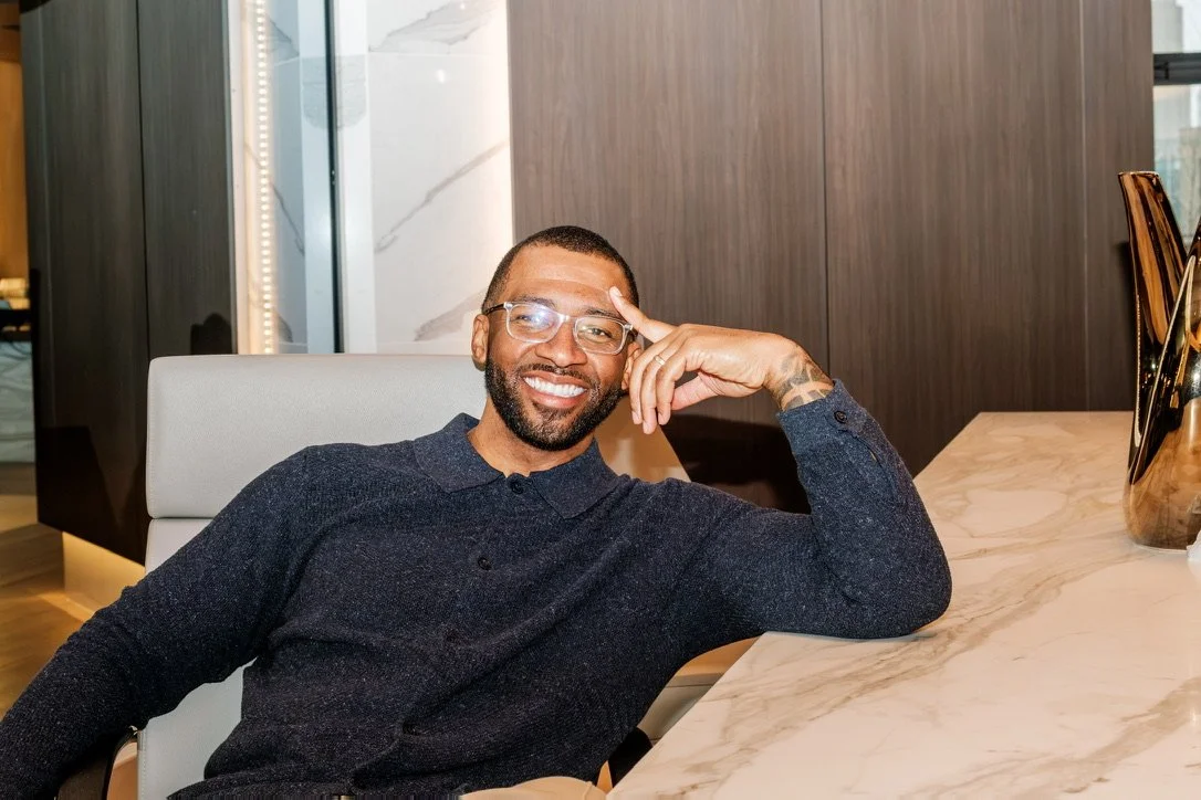 Chris Goode smiling, wearing glasses, sitting at a desk in a modern office.