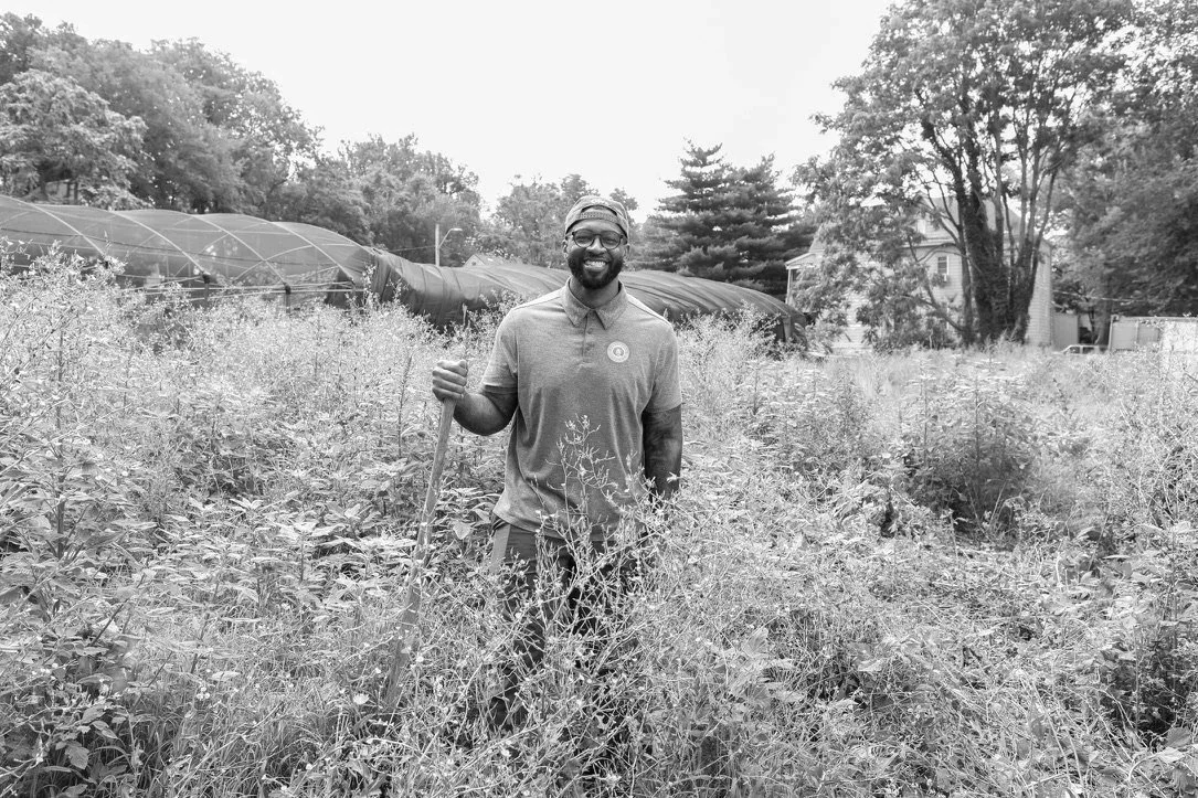 Chris Goode standing in a garden with tall plants, holding a gardening tool, surrounded by trees and a greenhouse in the background.
