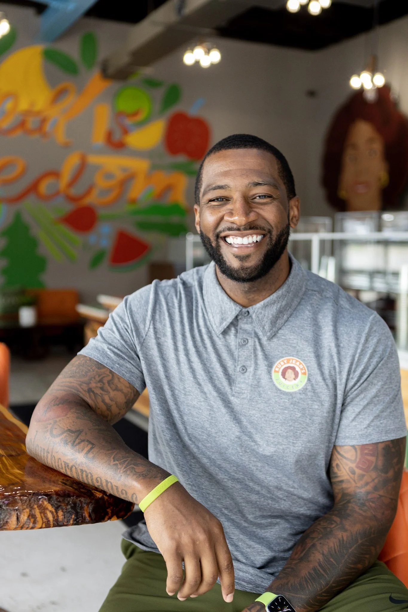 Chris Goode wearing a gray polo shirt, sitting at Ruby Jean's Juicery with colorful wall art in the background.