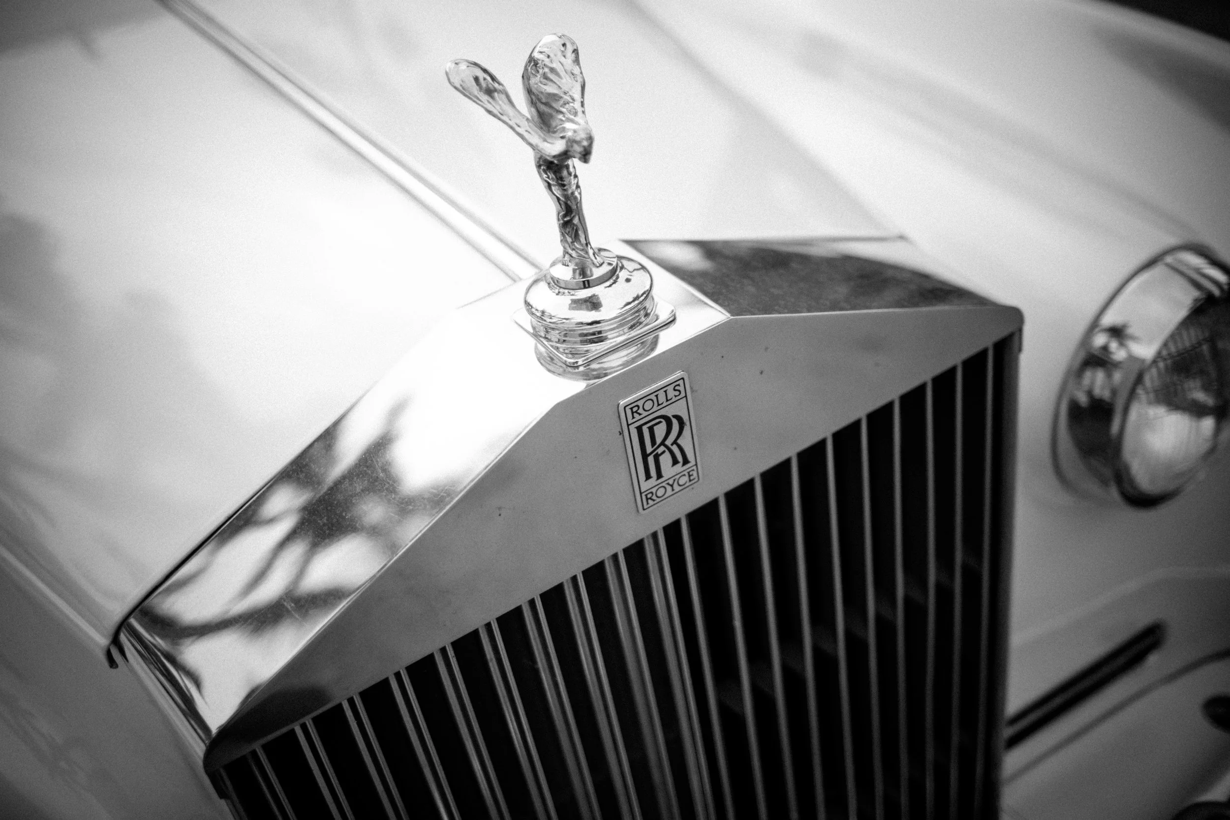 Frontal de un coche clásico Rolls Royce en blanco y negro, mostrando la capota con la ornamentación de la figura de un pato en la parte superior. Se observan detalles de la parrilla y el emblema con las letras 'RR' en la parte frontal.
