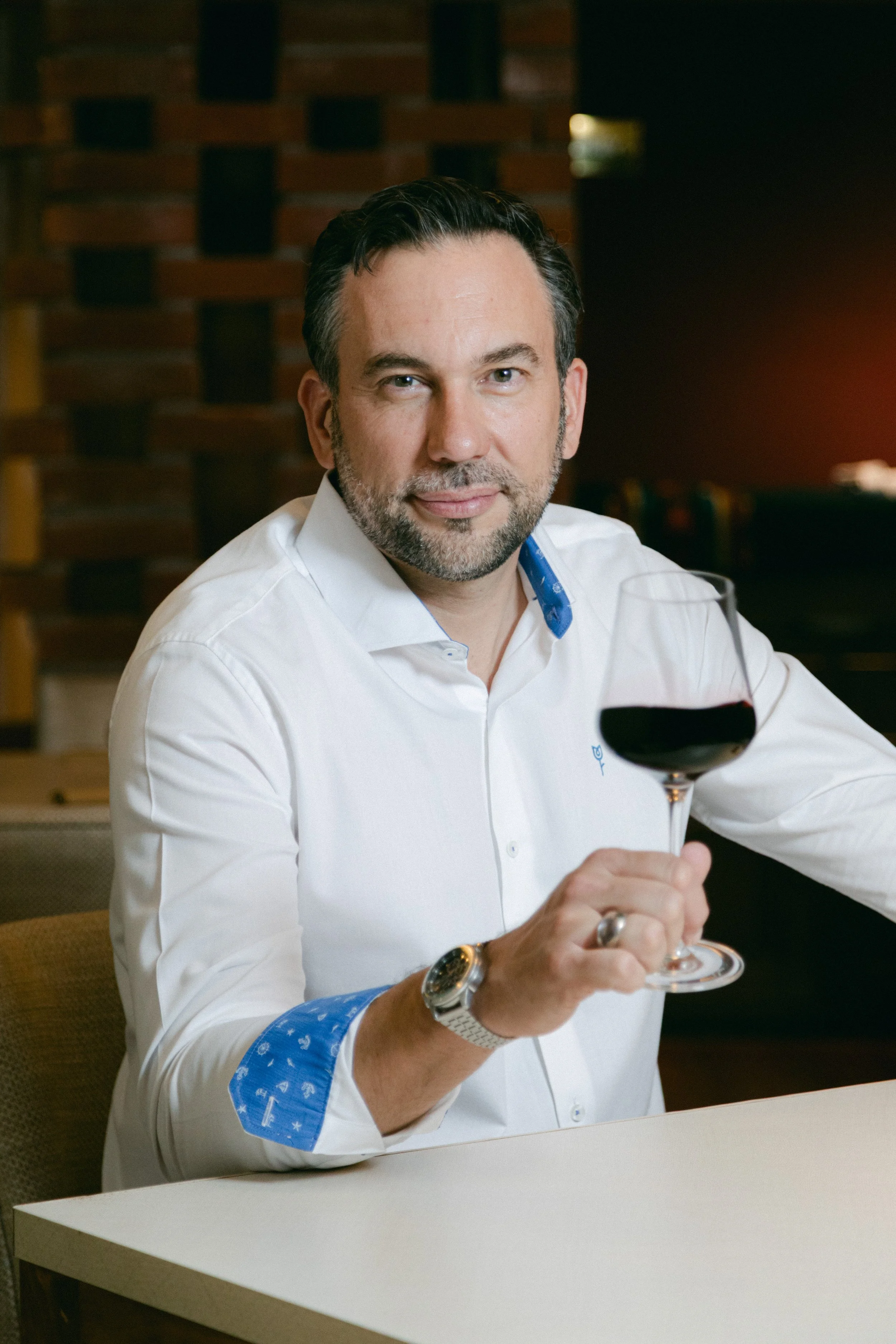 Hombre con barba, camisa blanca con detalles azules en las puños, sentado en una mesa, sosteniendo una copa de vino tinto en un ambiente de restaurante o bar.