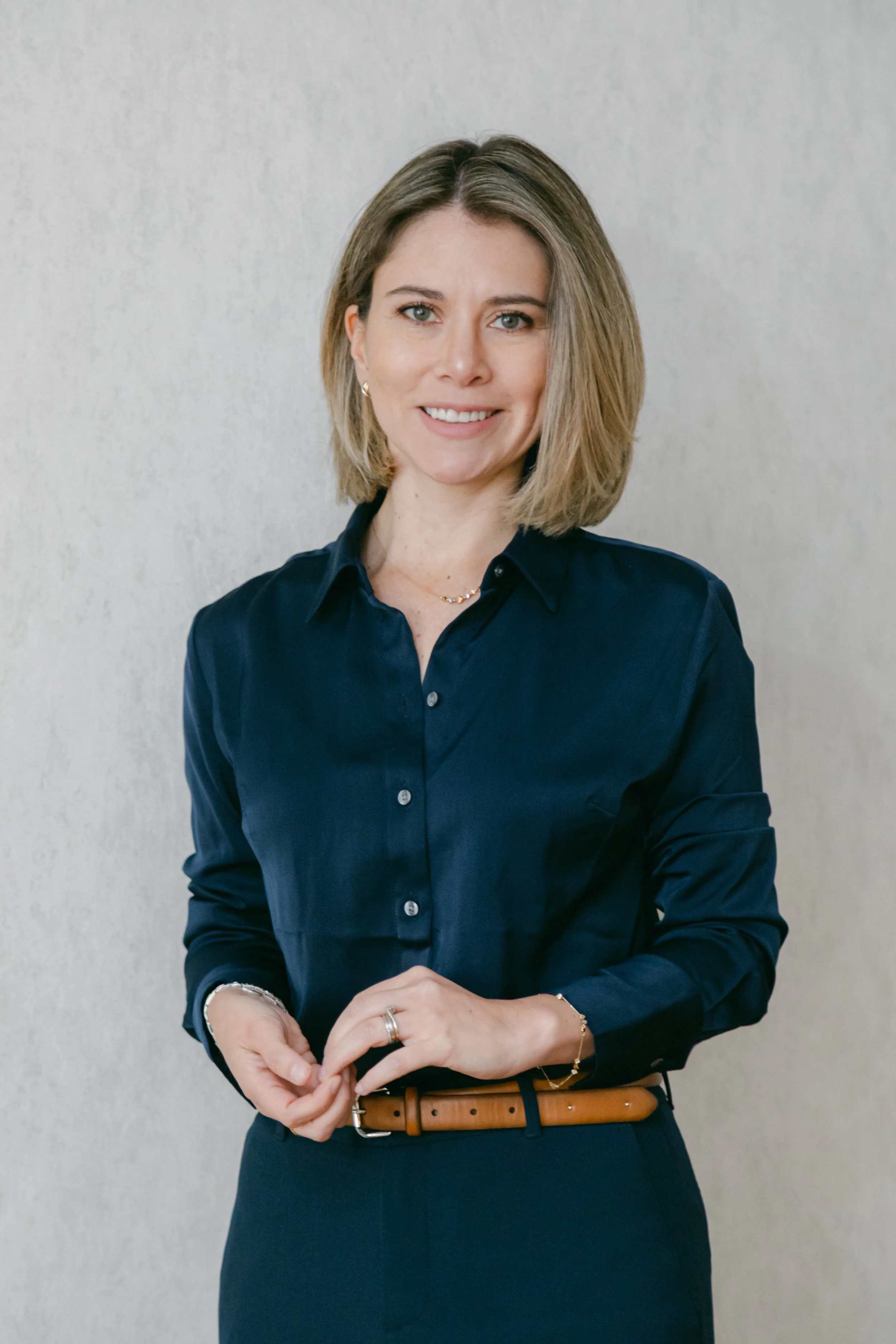 Mujer con cabello corto y rubio, vestida con blusa azul marino, sonriendo, en un fondo gris claro.