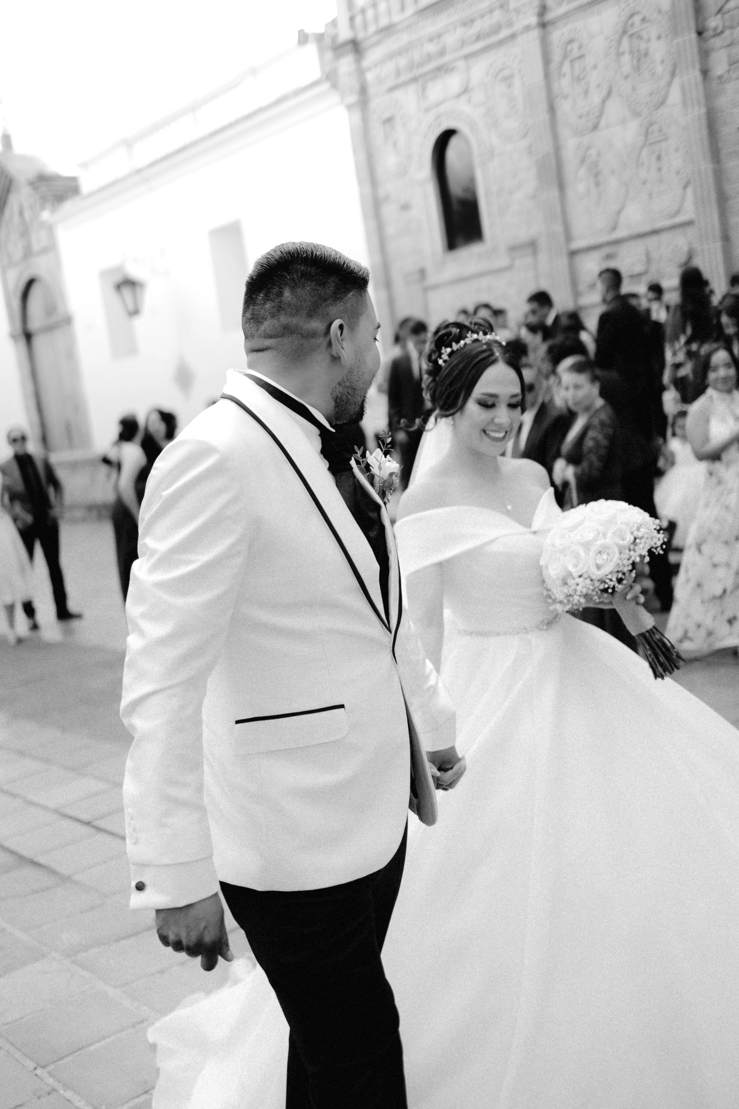Pareja de novios en una boda, la novia con vestido blanco y ramo de flores, el novio con saco blanco y corbata negra, caminando juntos entre invitados en una iglesia antigua.