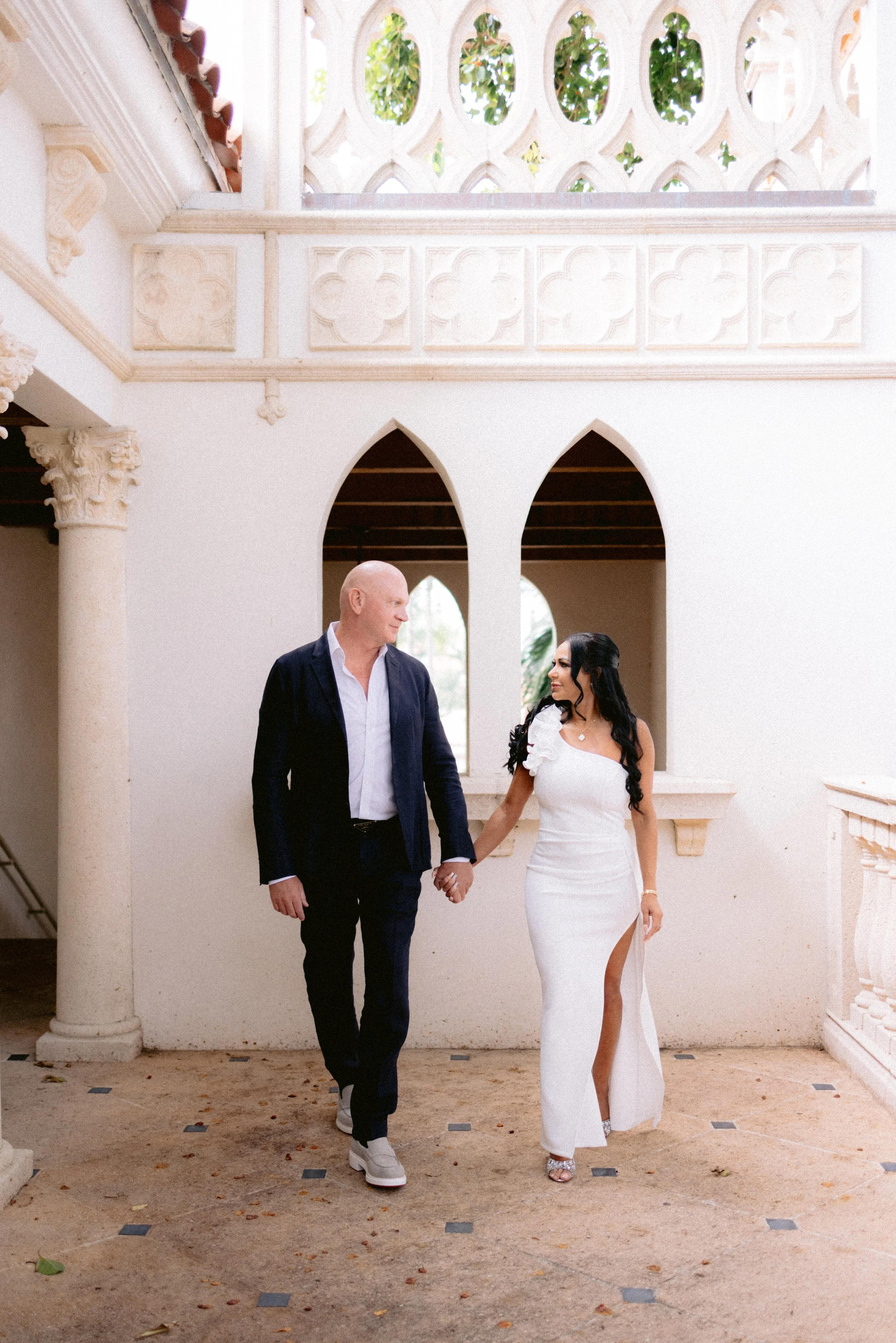 Una pareja, una mujer vestida con un vestido blanco con abertura en la pierna y un hombre con traje oscuro, caminando juntos por un espacio con arquitectura clásica, arcos y columnas, en un lugar luminoso y decorativo.