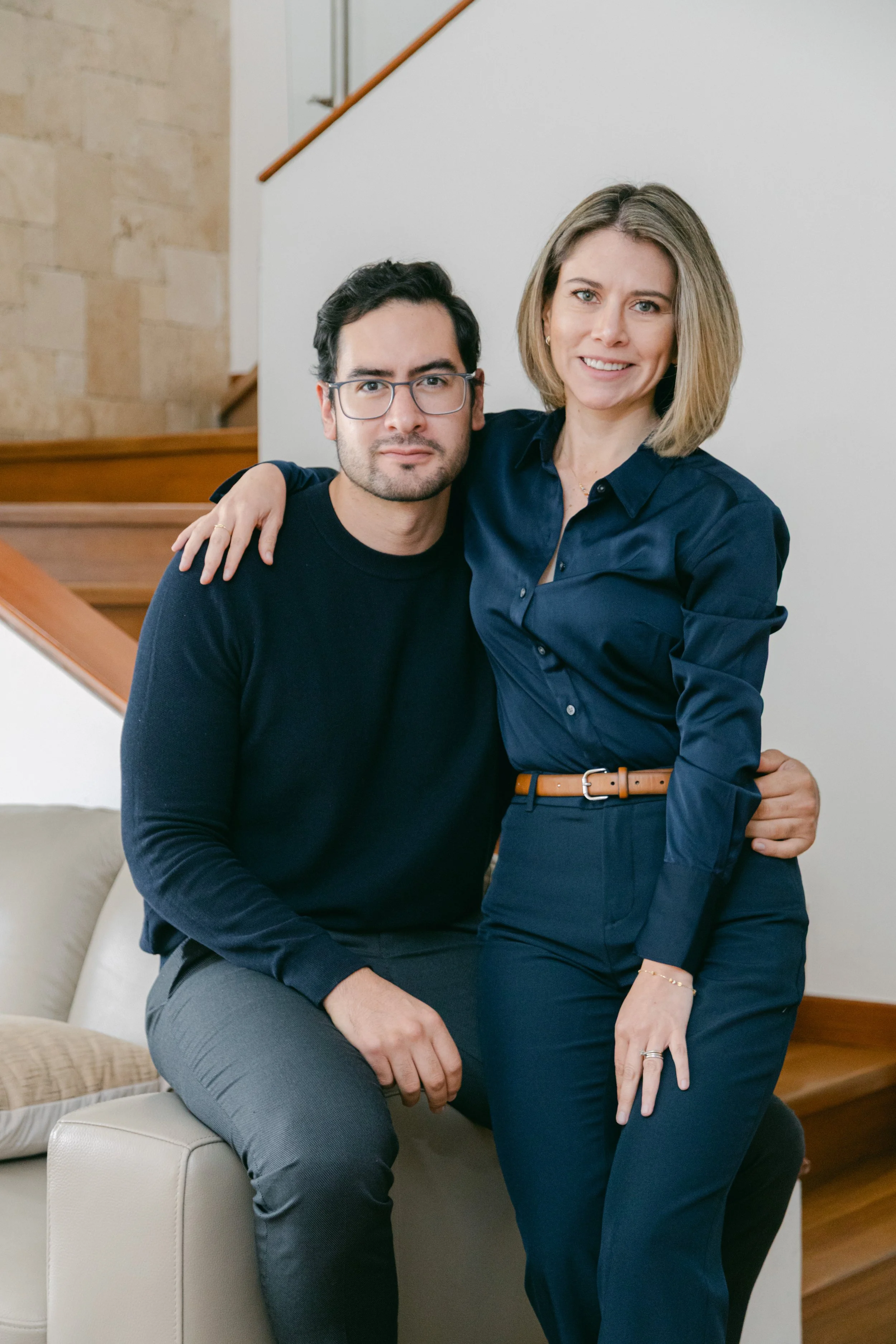 una pareja en un entorno interior, la mujer con blusa azul y pantalones a la moda, el hombre con gafas y suéter oscuro, ambos posando juntos en un ambiente hogareño