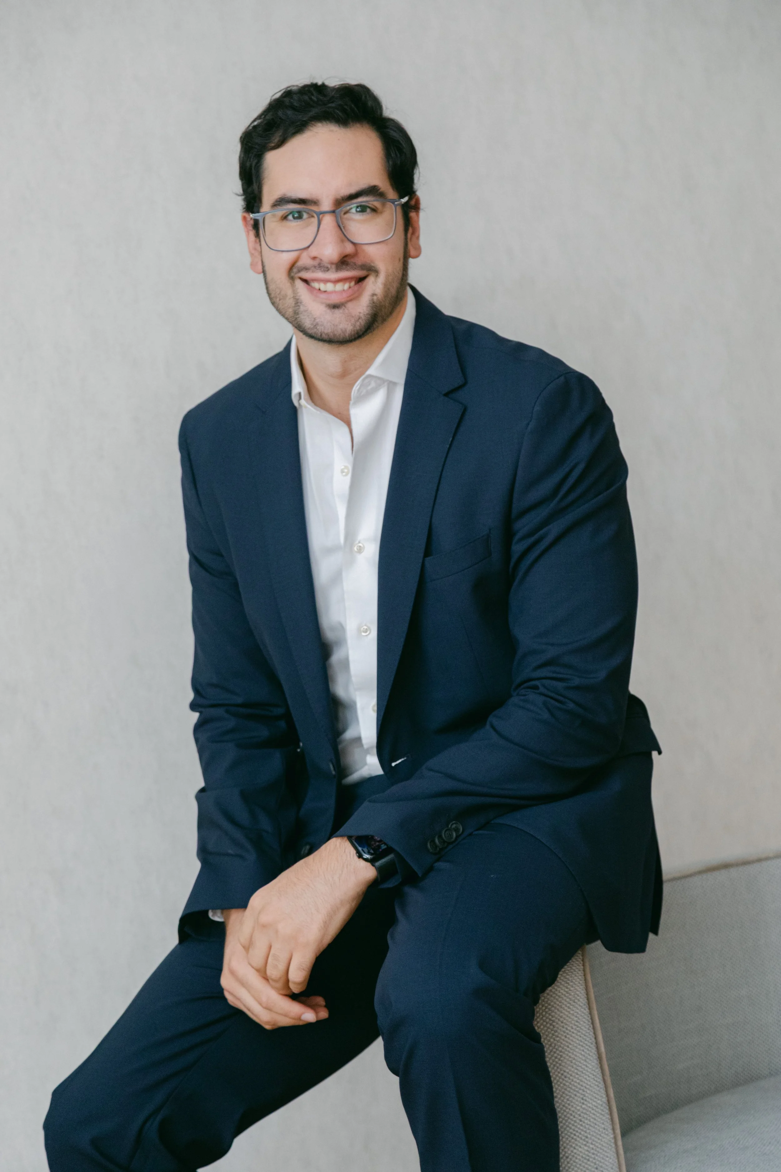 Un hombre sonriente con gafas, vestido con traje azul y camisa blanca, sentado en una silla con fondo gris.