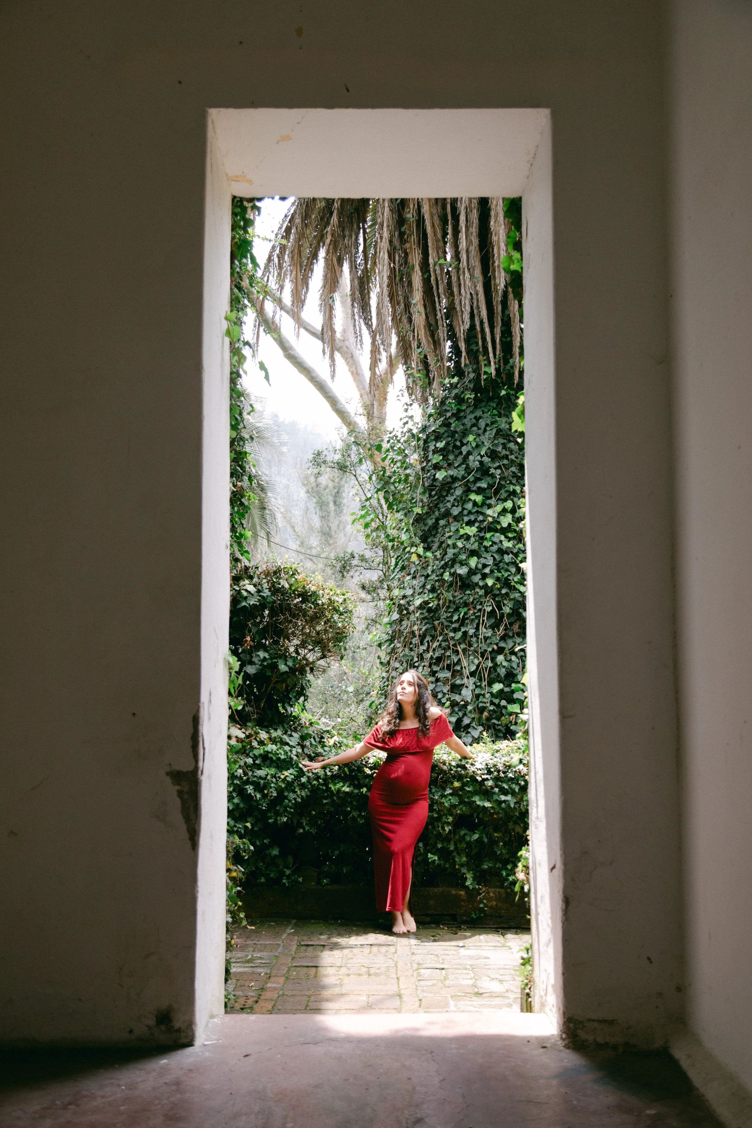 Mujer con vestido rojo de pie en una terraza rodeada de vegetación, vista desde el interior de un edificio a través de una puerta