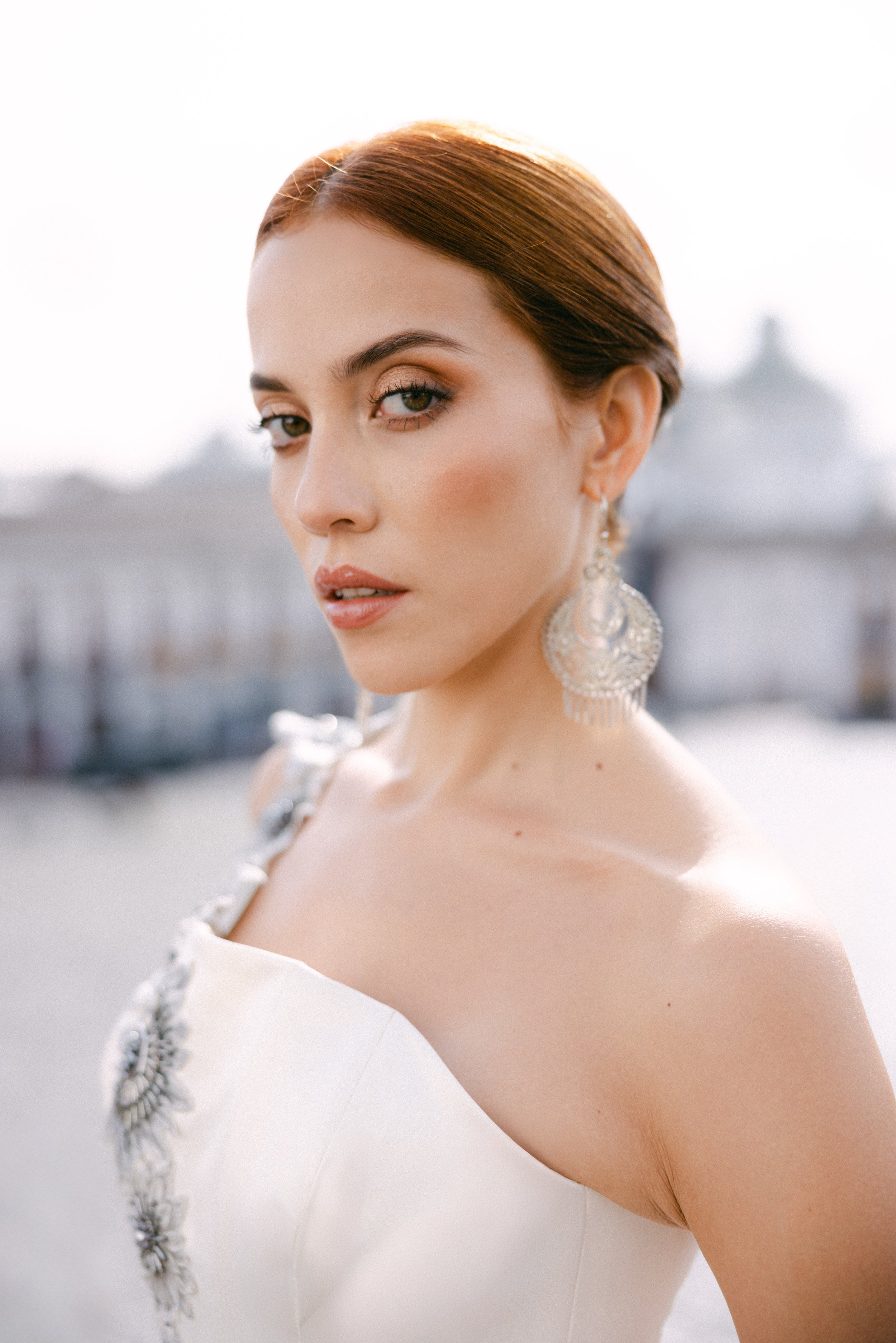 Retrato de una mujer con peinado en forma de raya y pendientes grandes, vestida con un vestido blanco con detalles plateados, al aire libre en un día soleado.