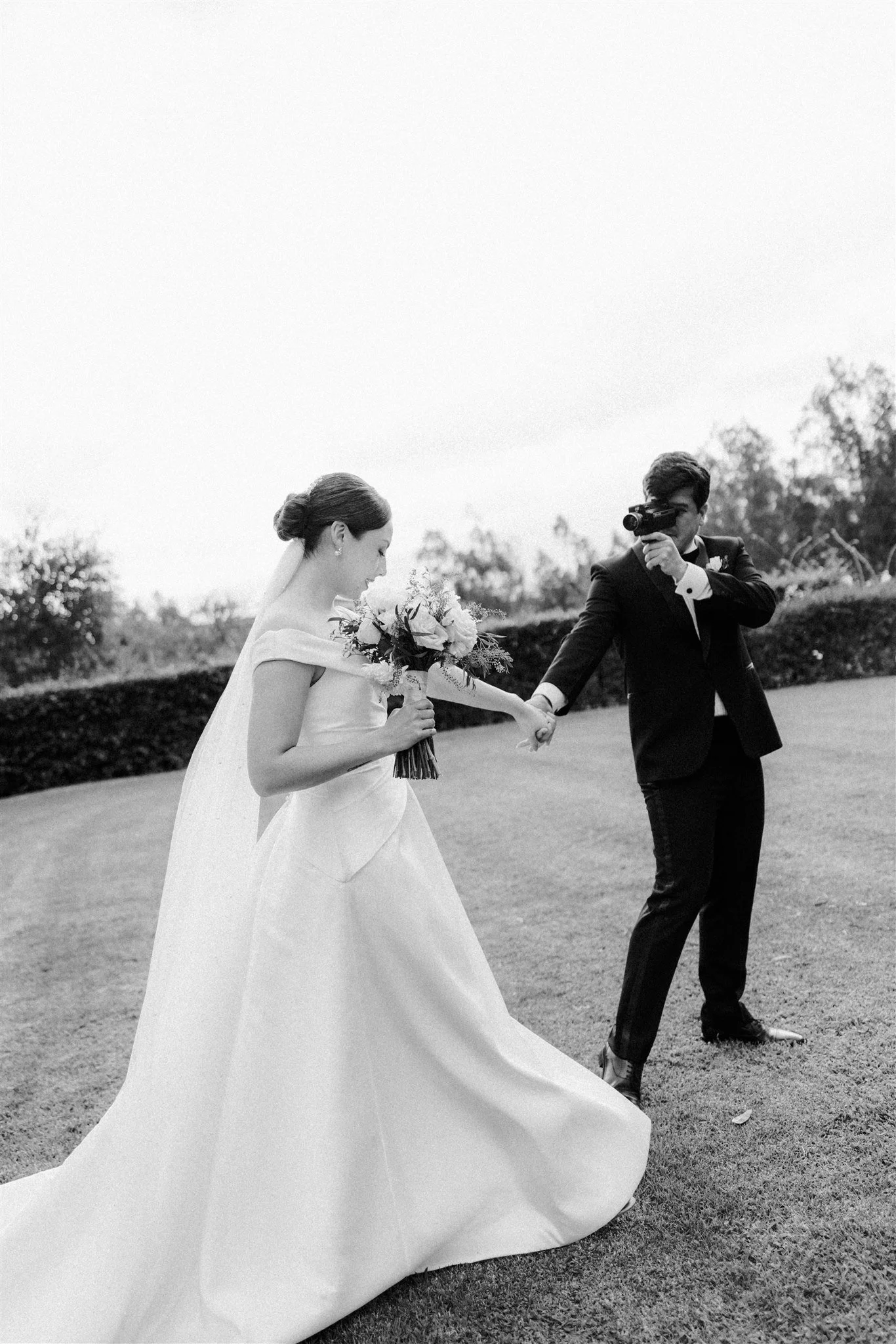 Pareja de novios en una boda, la novia sostiene un ramo de flores y el novio apunta con una cámara fotográfica, en un entorno exterior.