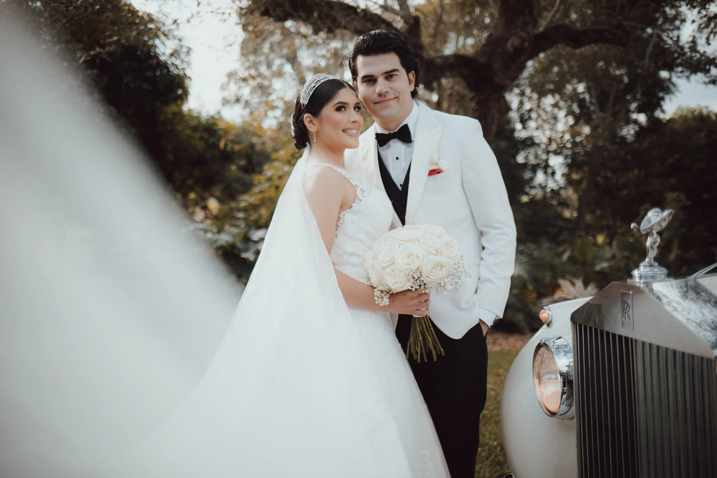 Una pareja de recién casados posa juntos al aire libre, con un árbol grande en el fondo y un coche clásico blanco en primer plano.