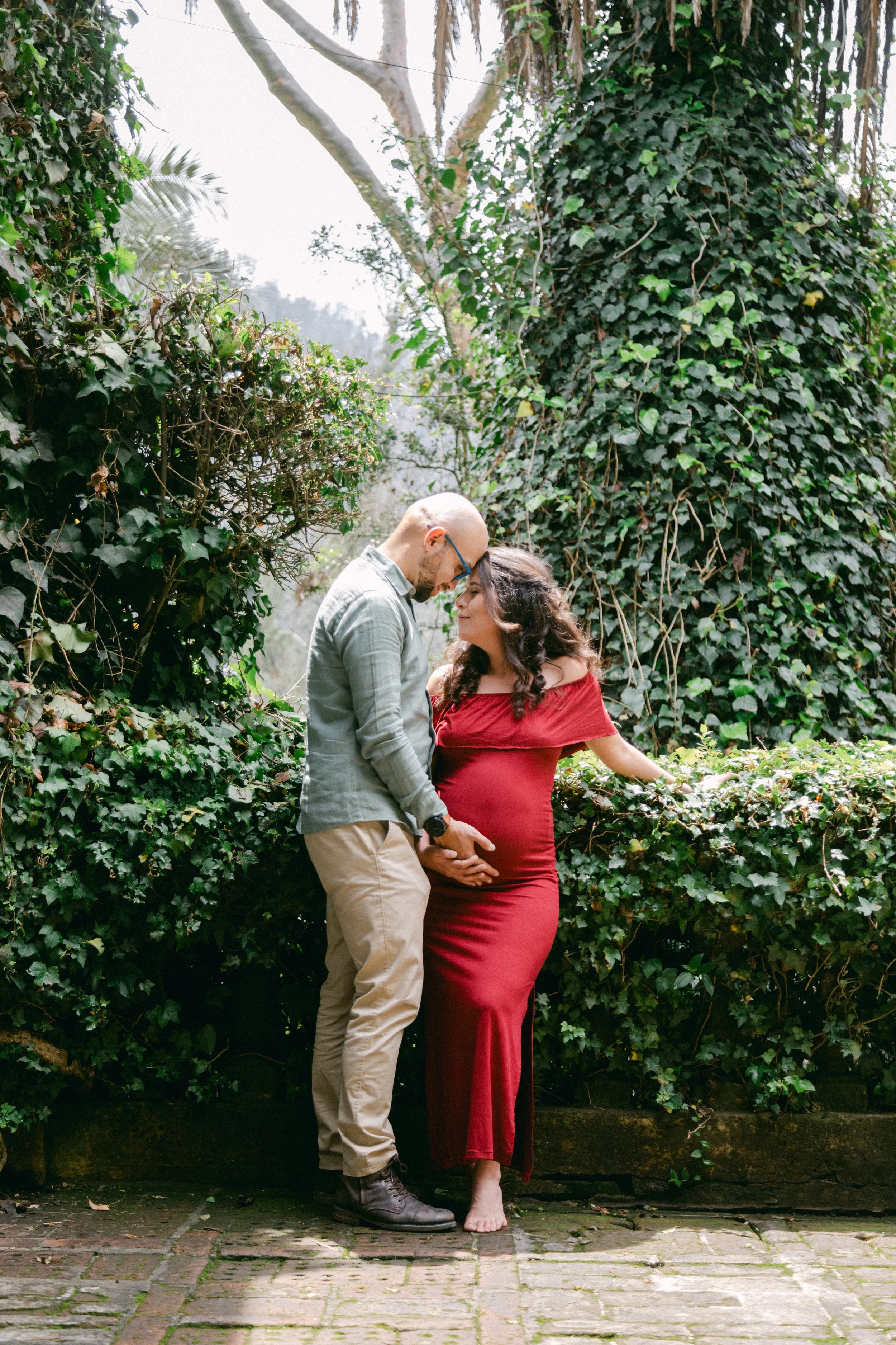 Una pareja en un jardín con mucha vegetación, se ven de frente, tocándose la panza, la mujer embarazada lleva un vestido rojo y el hombre lleva una camisa gris y pantalones beige.