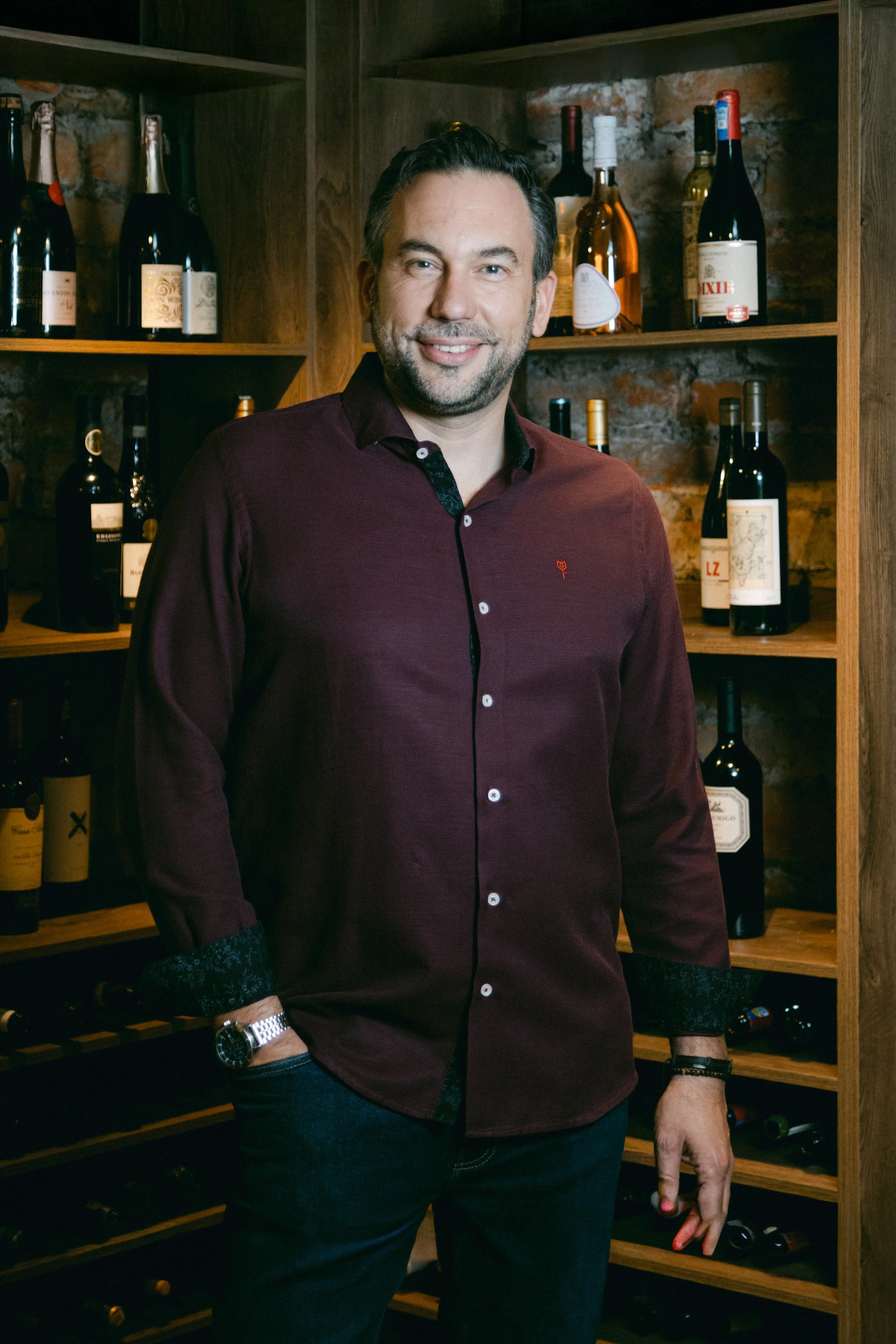 Hombre sonriendo en una cava de vinos, con estantes llenos de botellas de vino a su alrededor.