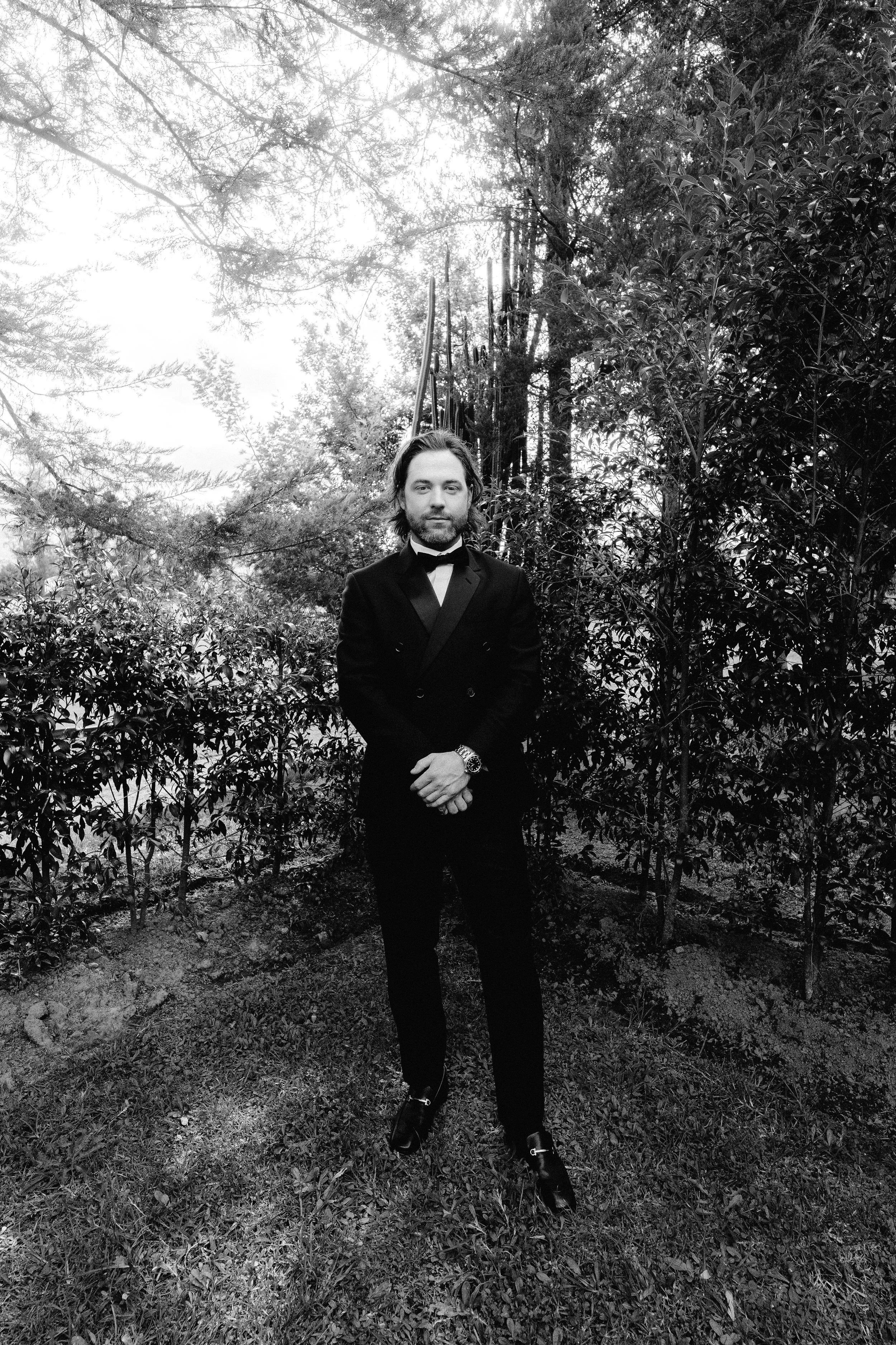 Hombre en traje formal posando en un jardín con árboles y arbustos.