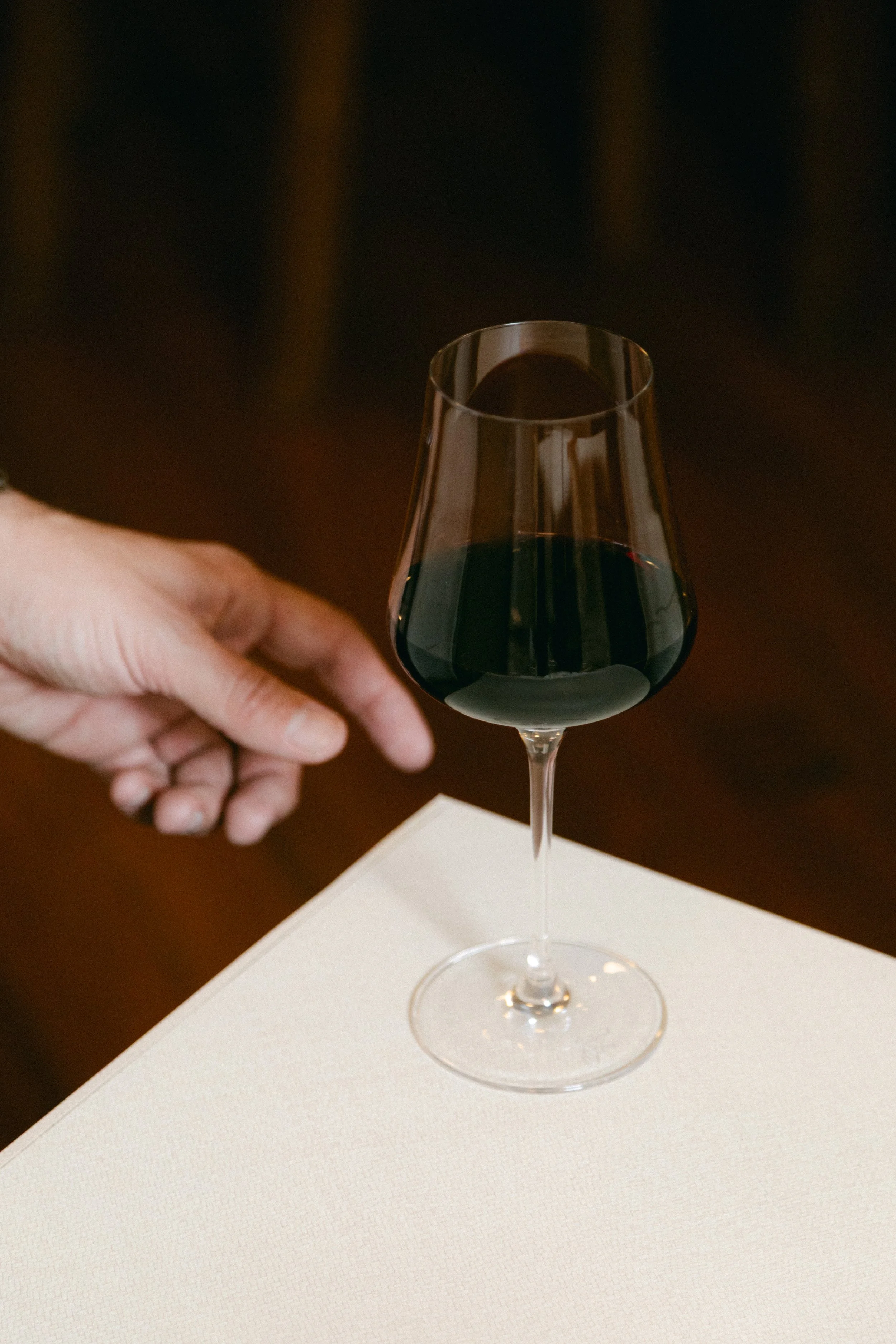 Una copa de vino tinto sobre una mesa blanca, con una mano a la izquierda que parece estar a punto de servirla.