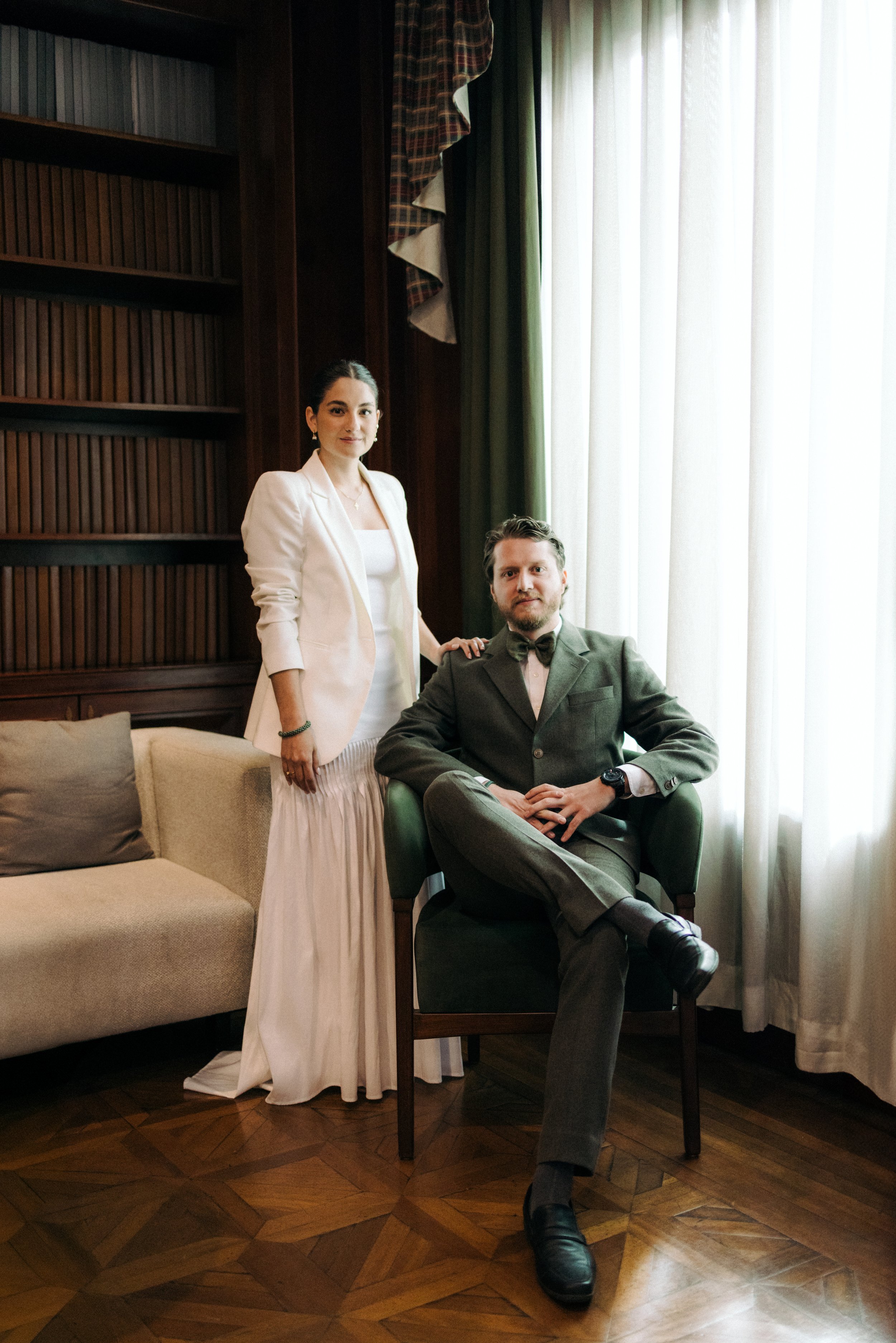 Una mujer de vestido blanco y saco blanco de pie junto a un hombre de traje sentado en una habitación con cortinas blancas y paredes de madera.