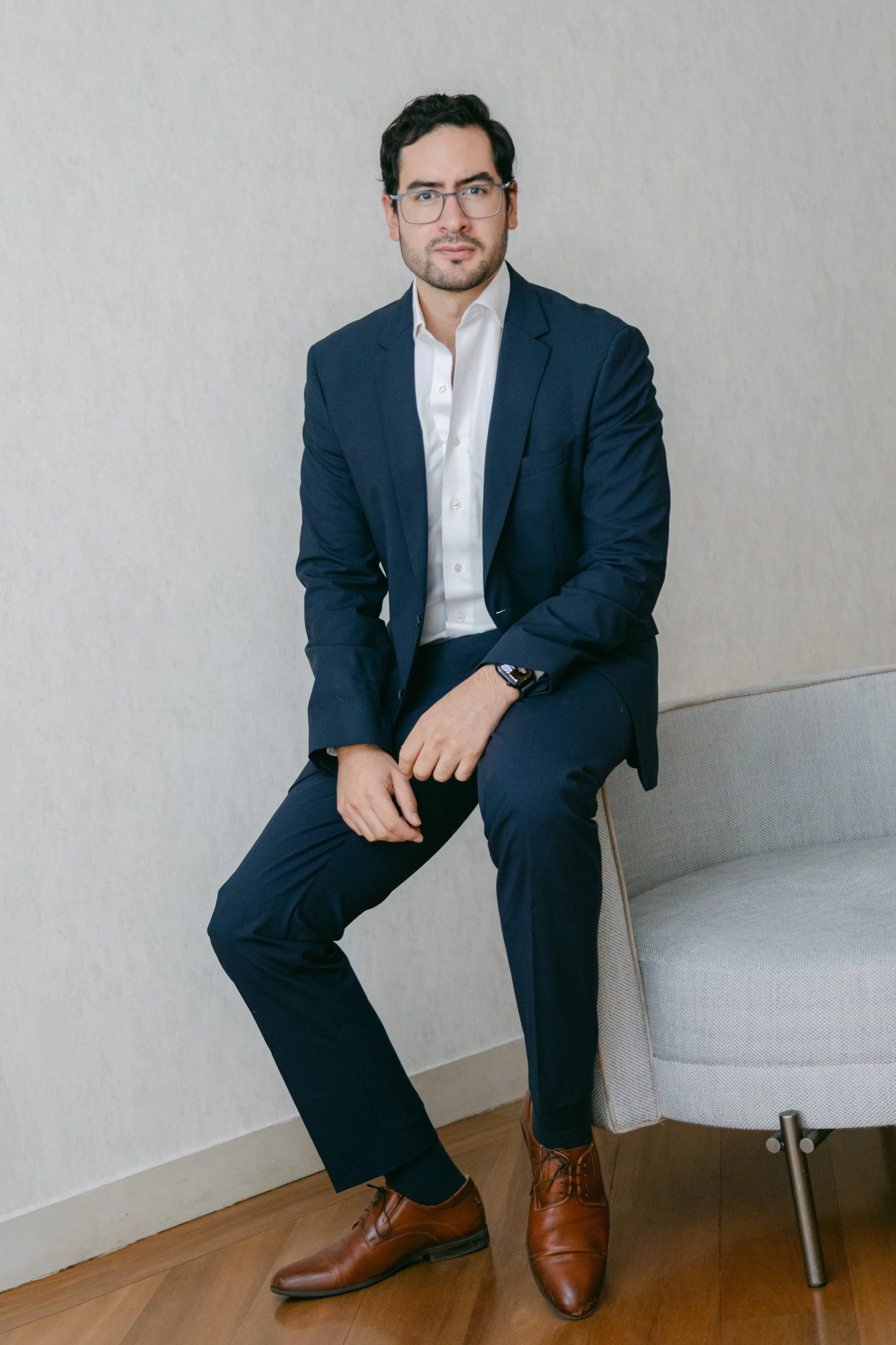 Hombre con gafas, traje azul, camisa blanca, sentado en una silla y miranda hacia la cámara.