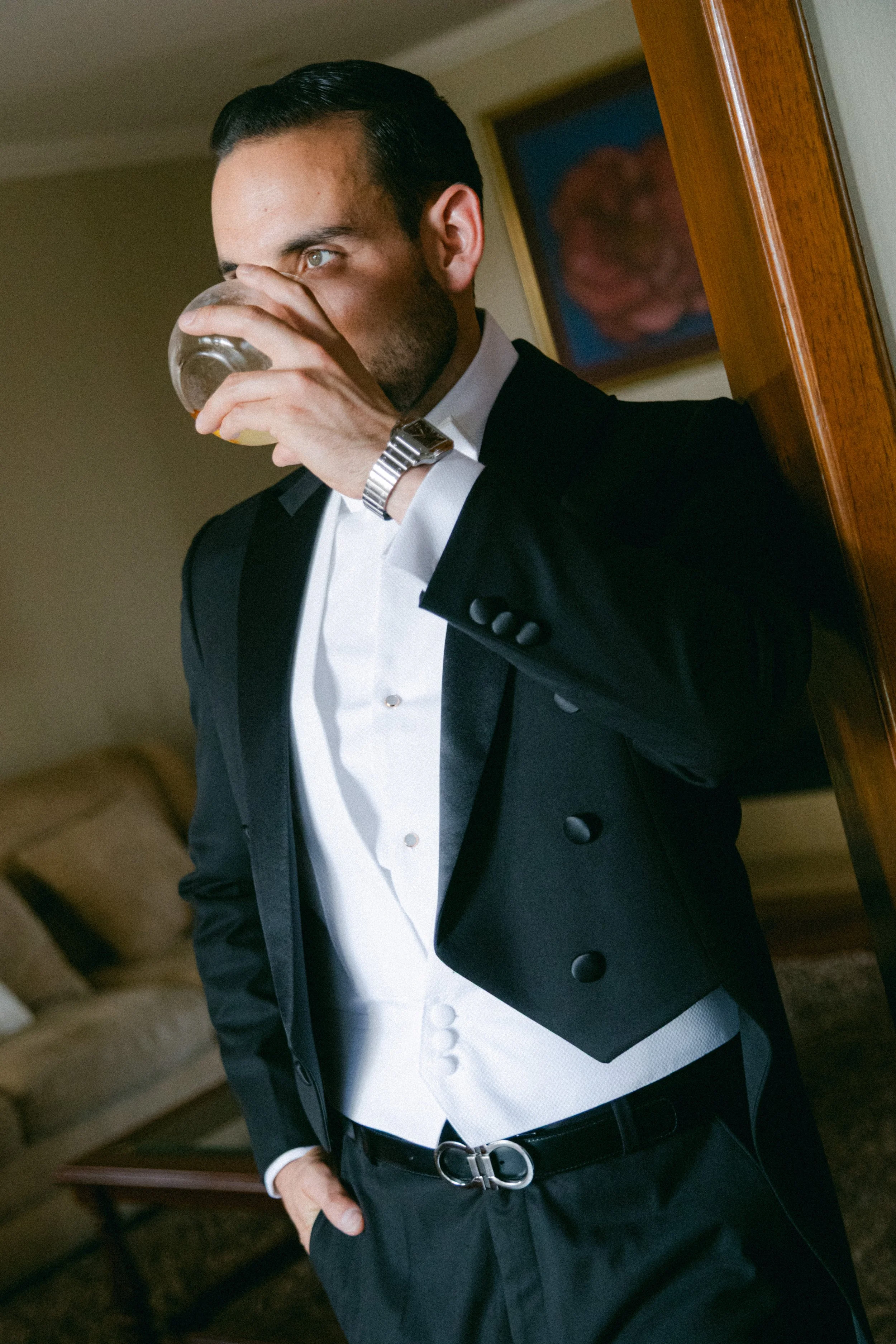 Hombre con ropa formal y reloj bebiendo de un vaso en una habitación elegante.