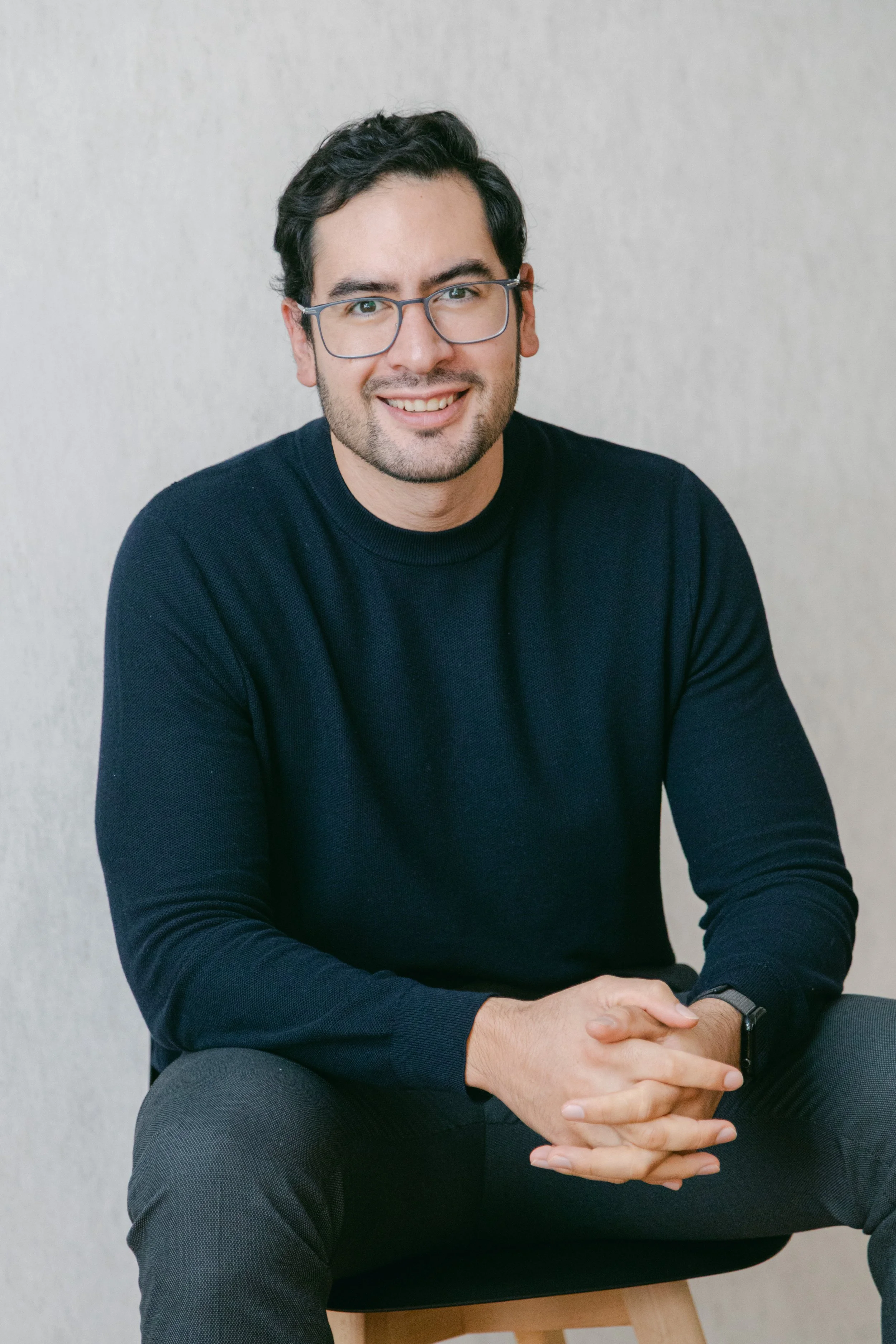 Hombre con gafas y barba, sonriendo, vestido con suéter negro, sentado en una silla de madera contra pared blanca.
