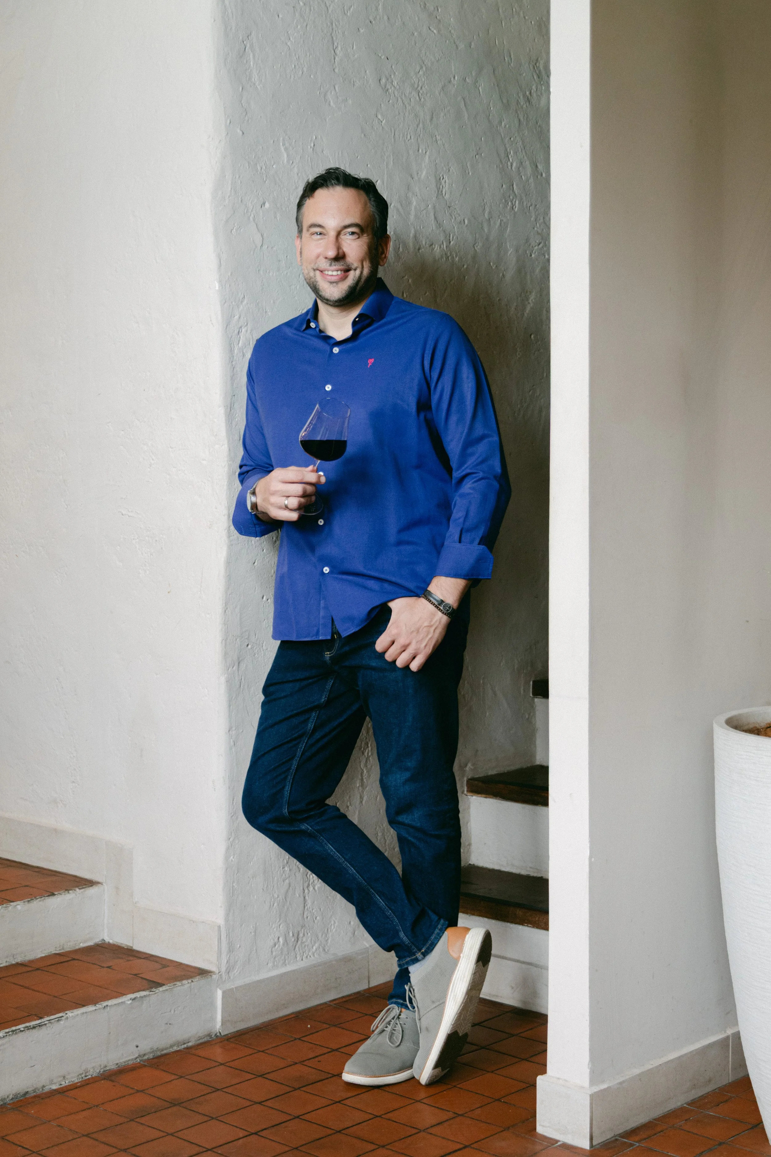 Hombre sonriendo, con barba y cabellera oscura, viste camiseta azul y jeans, sosteniendo una copa de vino tinto, en interior con paredes blancas y piso de azulejo.