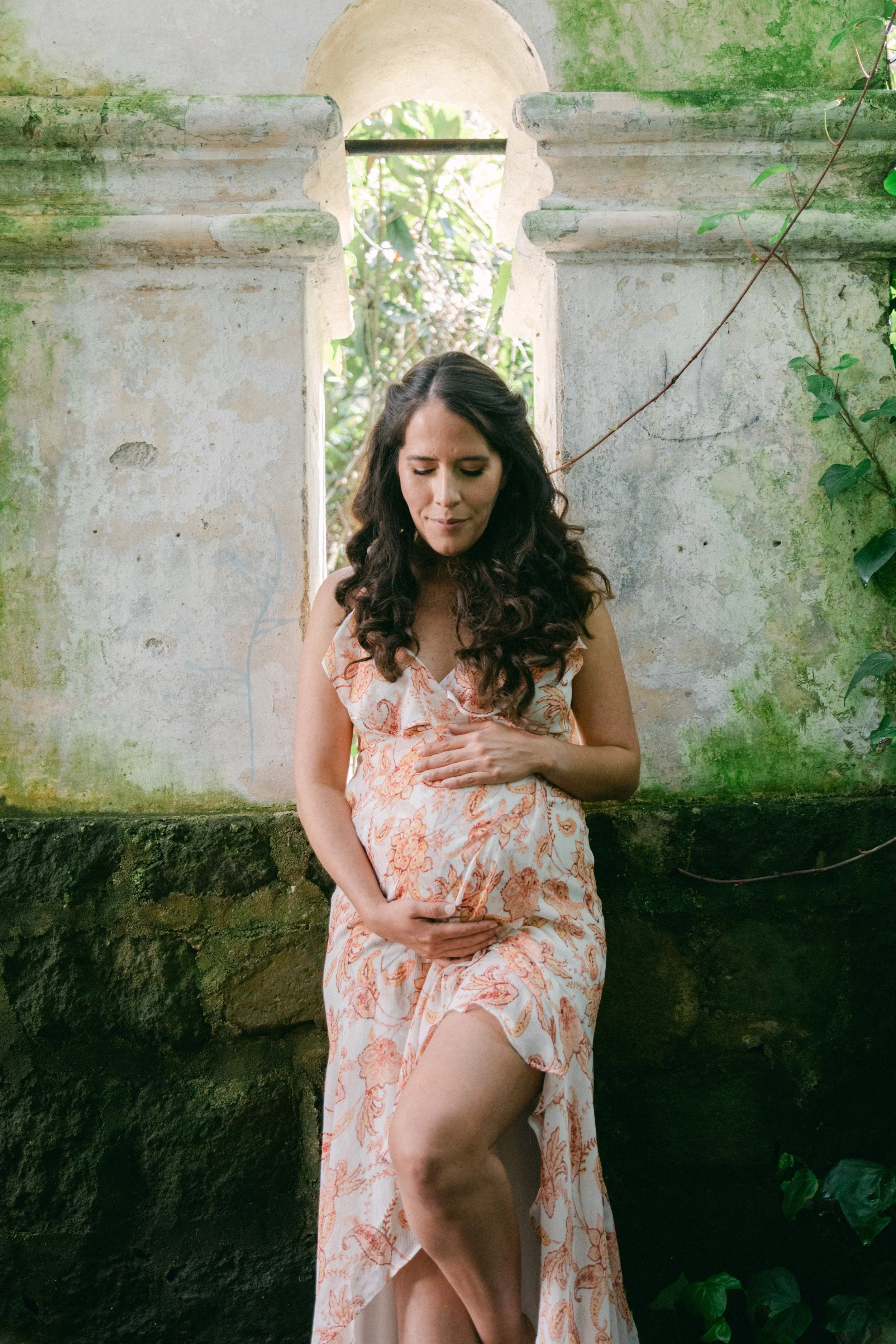 Mujer embarazada con vestido de estampado floral, posando en un entorno natural con paredes envejecidas y vegetación creciente.