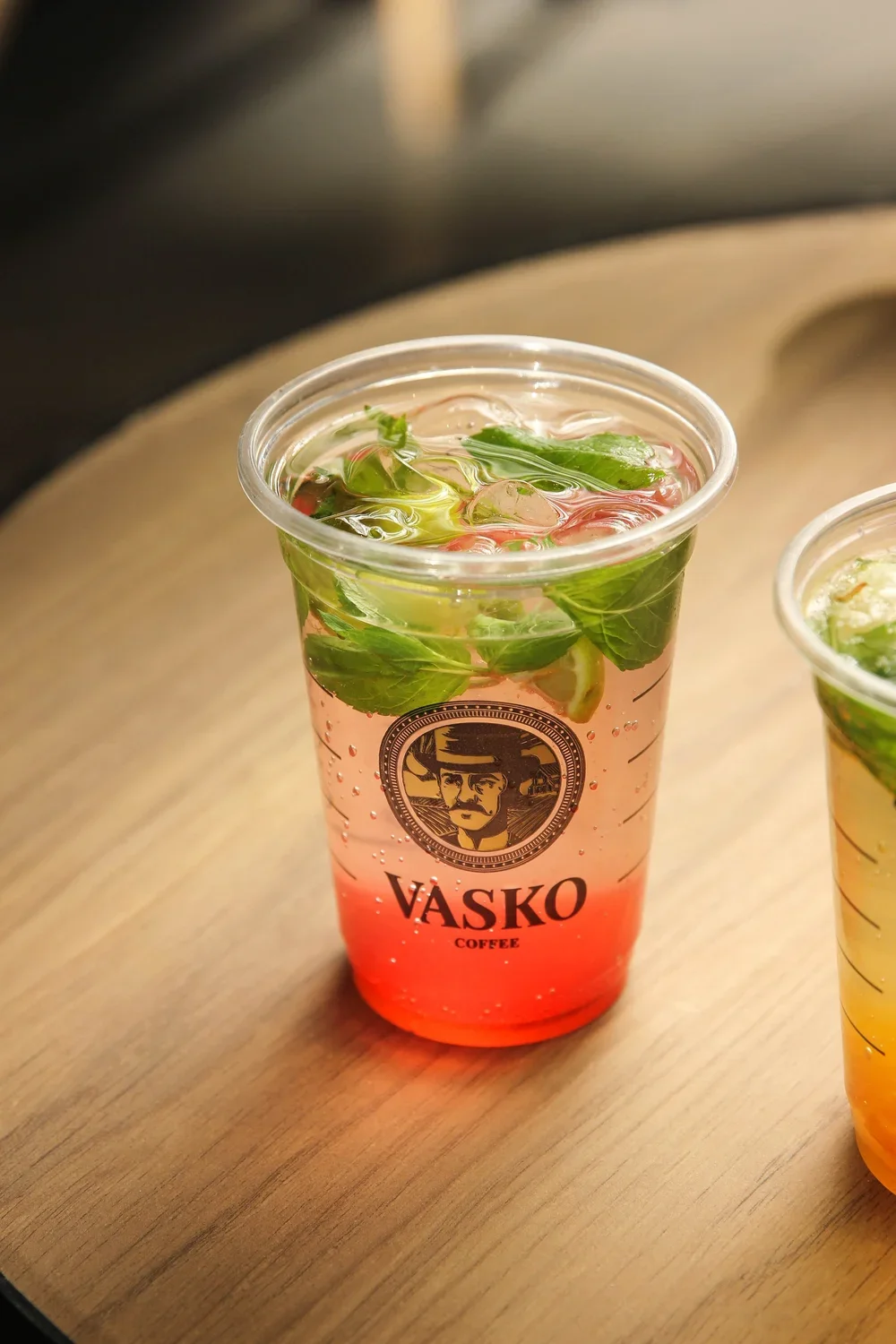A plastic cup from Väskö Coffee filled with a pinkish drink, ice, and fresh mint leaves on top, placed on a wooden surface.
