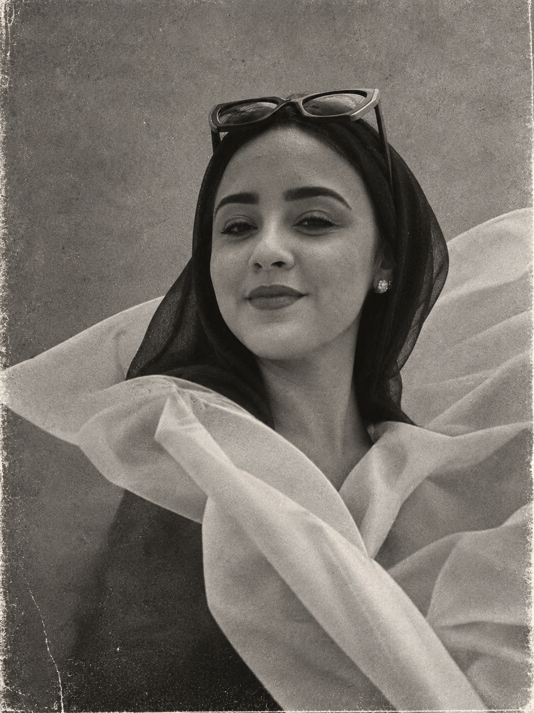 A woman wearing a headscarf and sunglasses on her head, smiling slightly, with earrings and a large, ruffled garment or accessory around her shoulders.