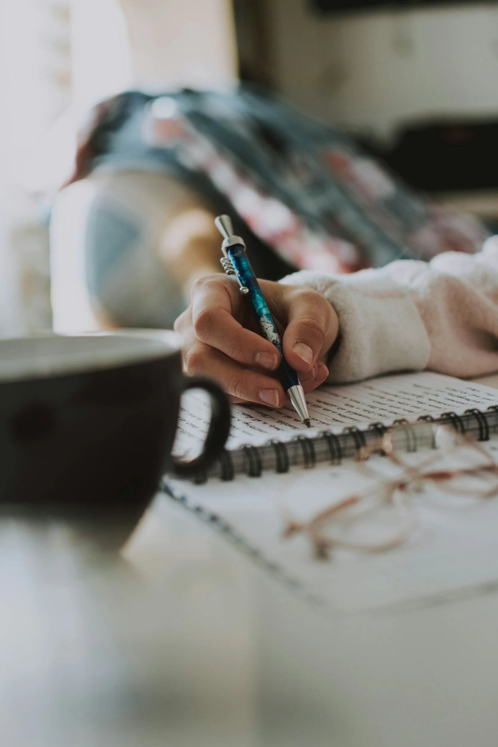 Close-up of a person writing in a notebook with a blue pen, with glasses and a black mug on the table.