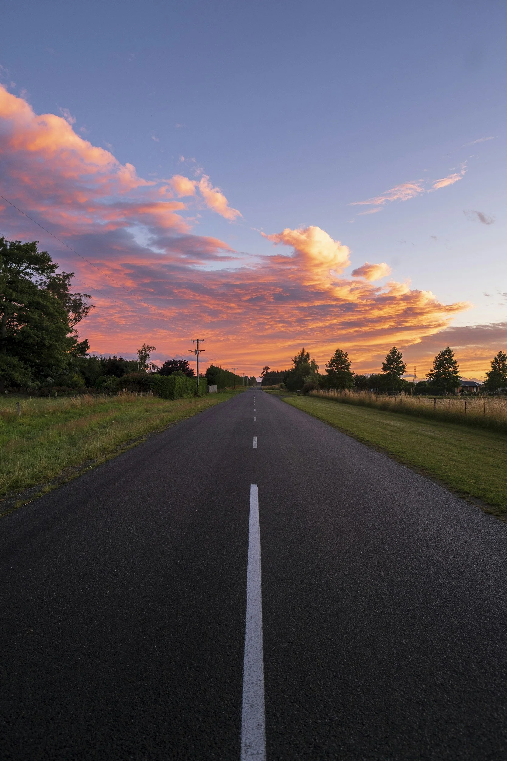 Long open road with white dashed lines, flanked by grassy fields and trees, under a colorful sky with pink and orange clouds at sunset.