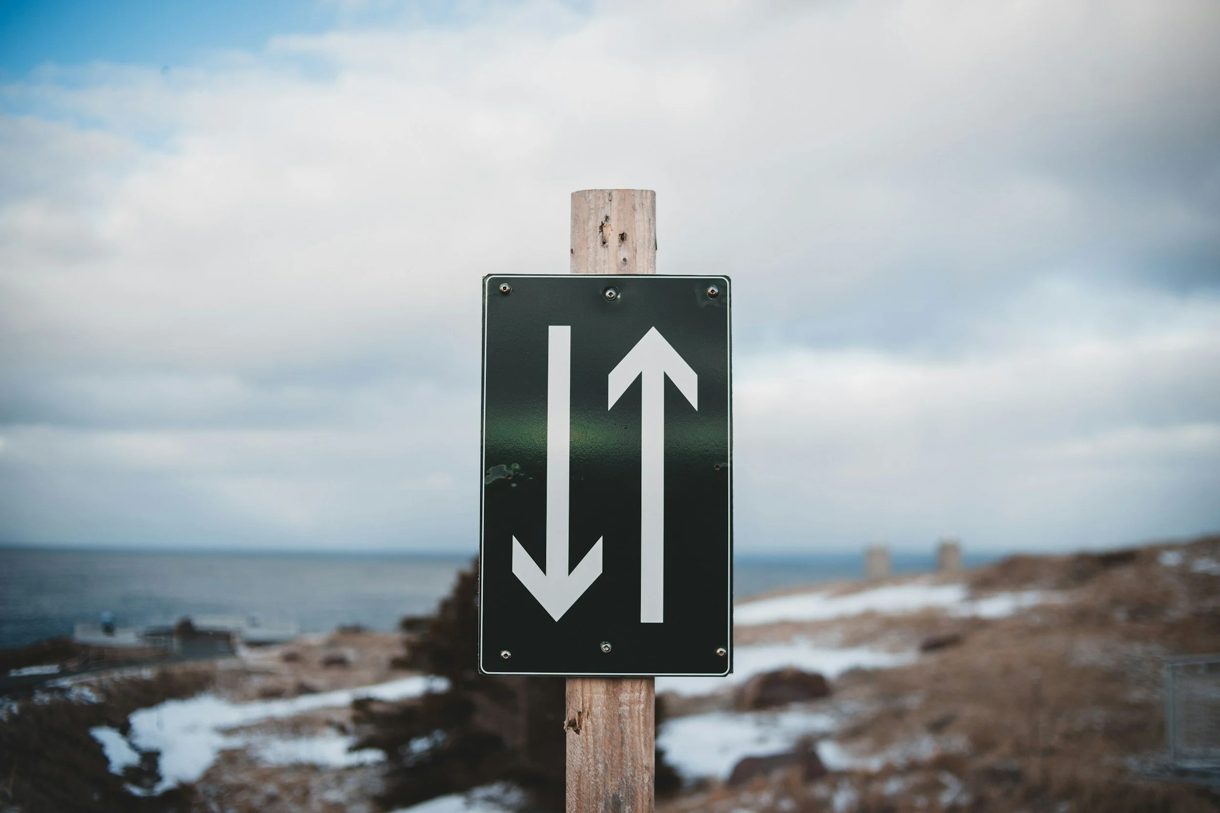 A directional sign on a wooden post with arrows pointing up and down, indicating two-way direction, near a coastal area with rocks, water, and a cloudy sky.