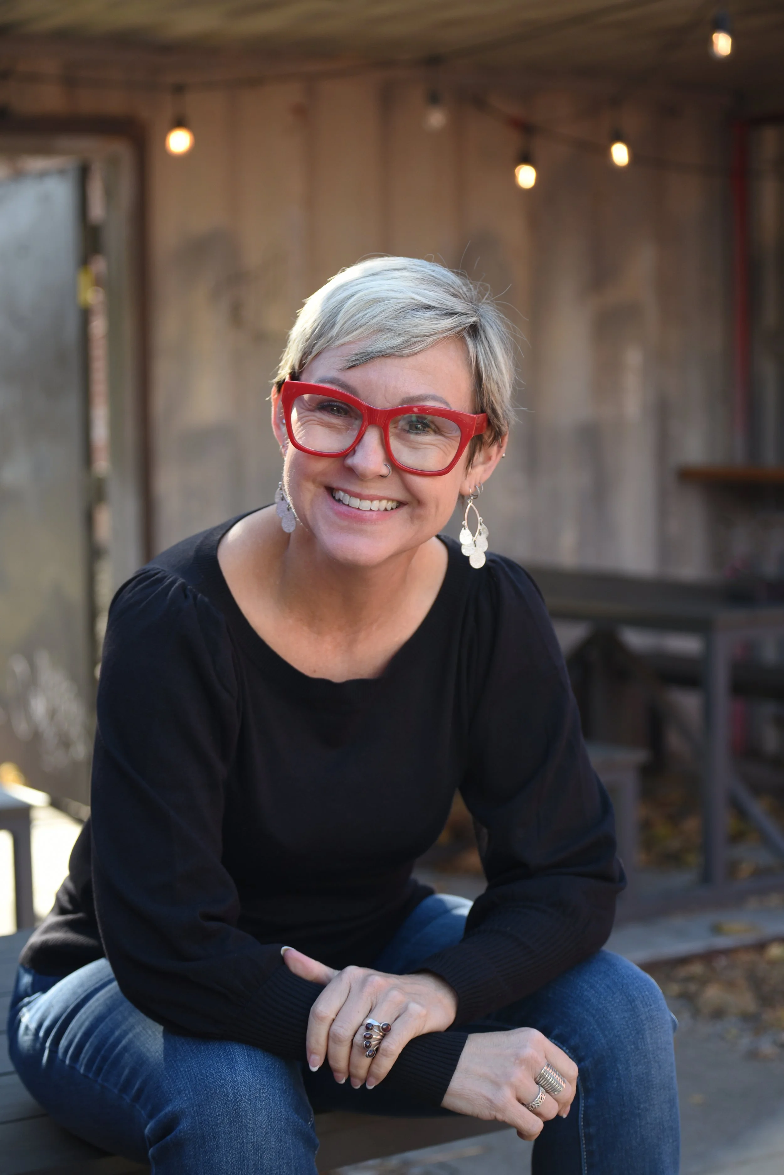 A woman with short blond hair and red glasses sitting outdoors, smiling at the camera, with a wooden structure and string lights in the background.