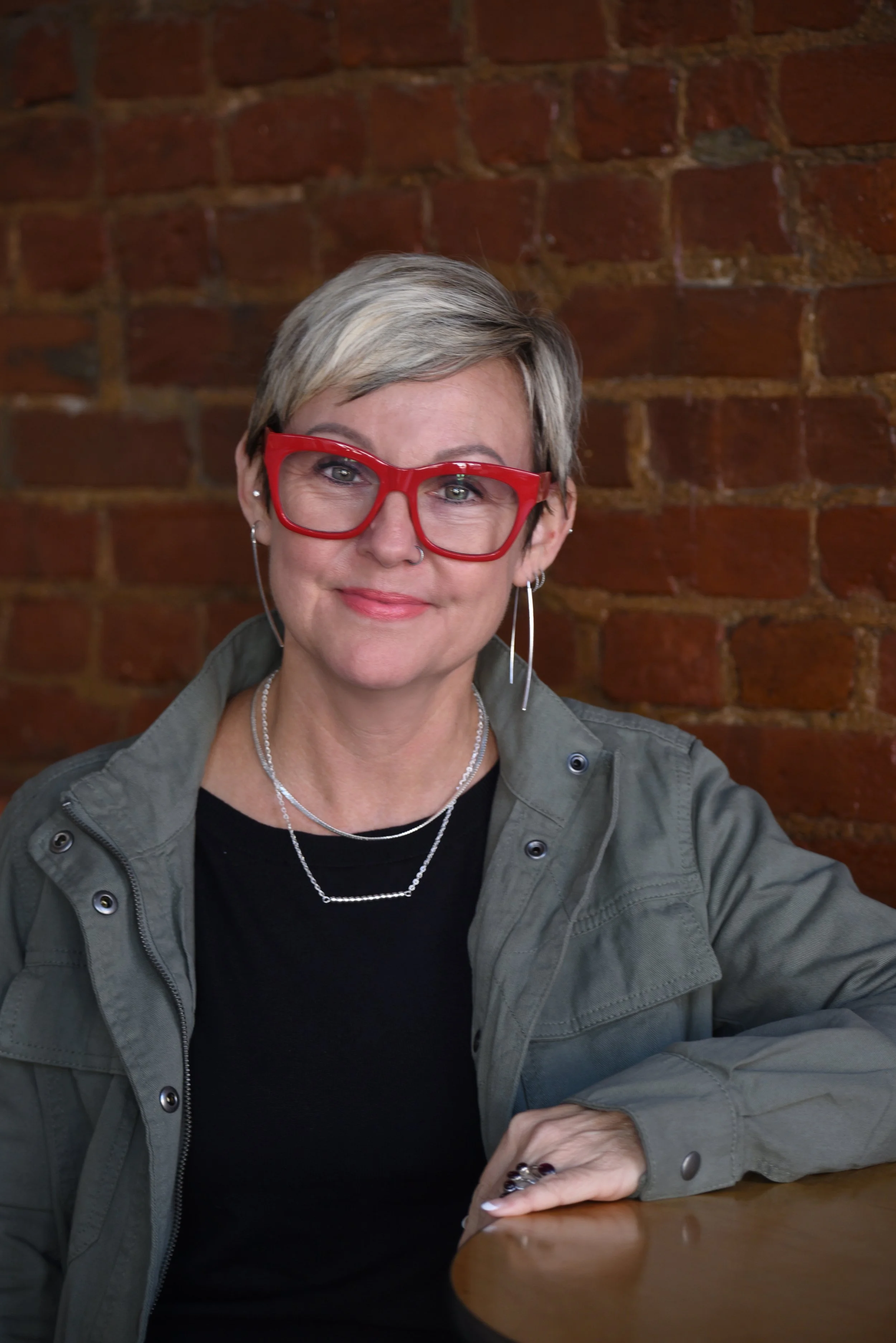 A woman with short blond hair wearing red glasses, silver jewelry, and a green jacket sitting at a table with a brick wall in the background.