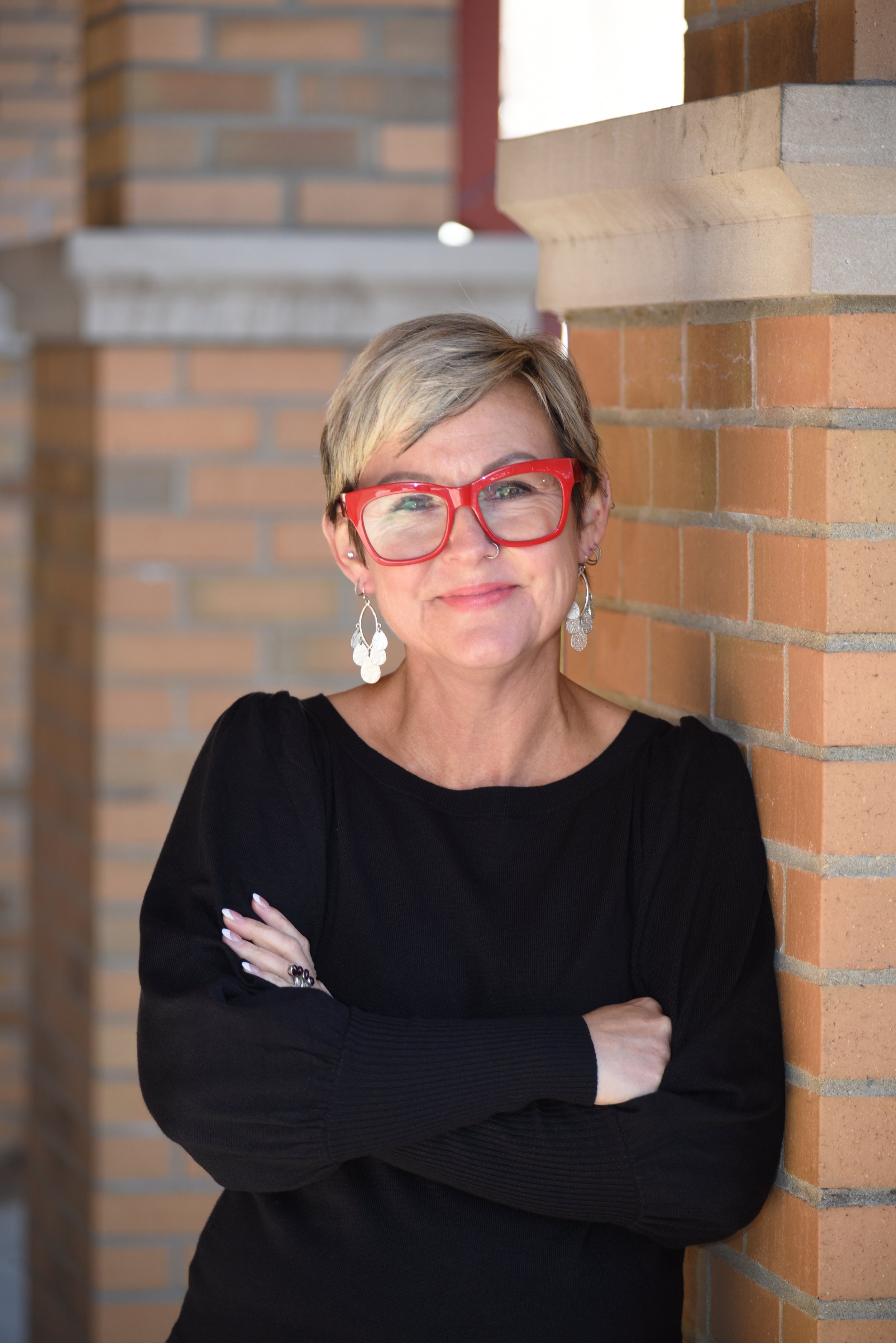 Portrait of a woman with short blond hair, red glasses, earrings, wearing a black top, leaning against a brick wall.