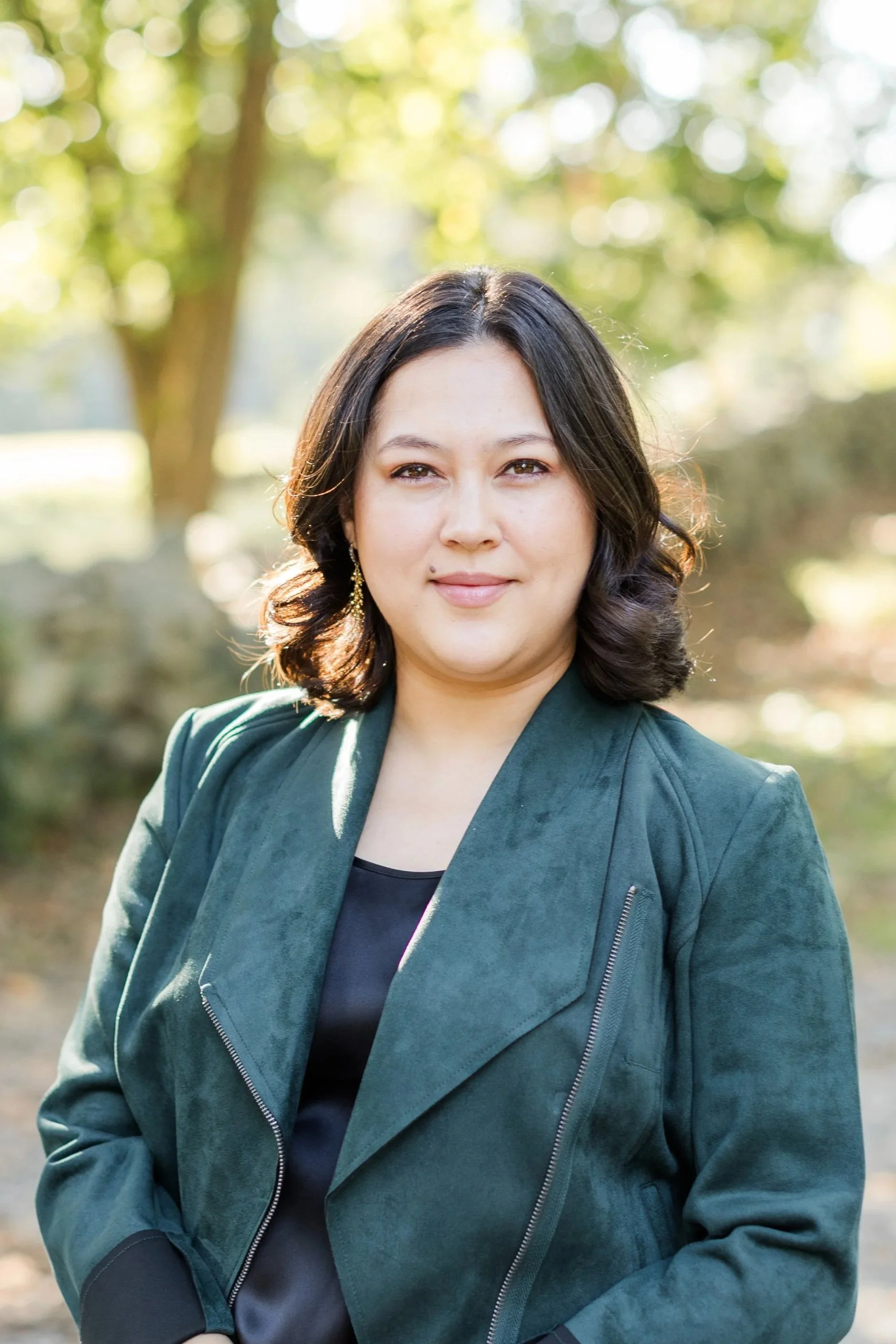 A woman with shoulder-length dark hair and light skin, wearing a dark green jacket and earrings, standing outdoors in a park with trees and sunlight in the background.