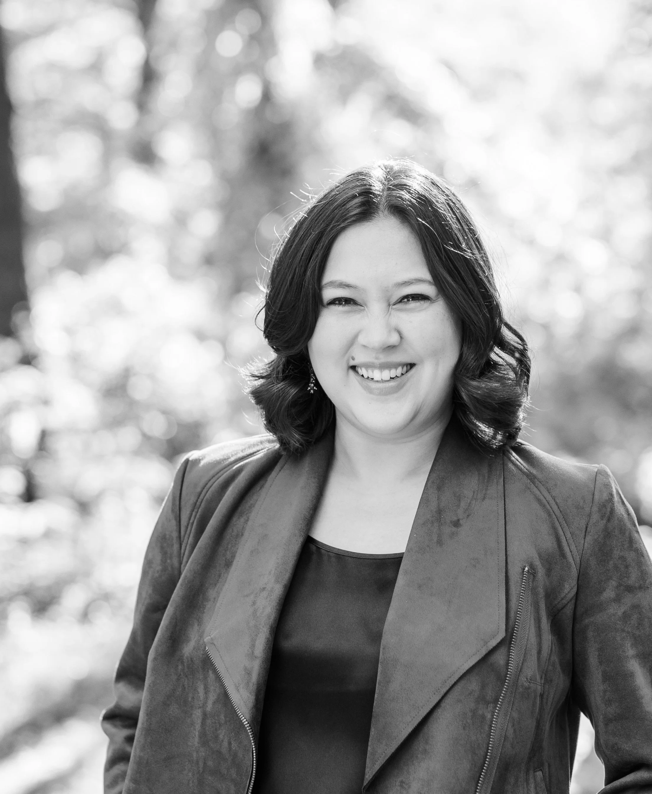 Black and white portrait of a woman smiling outdoors, with blurred trees in the background.