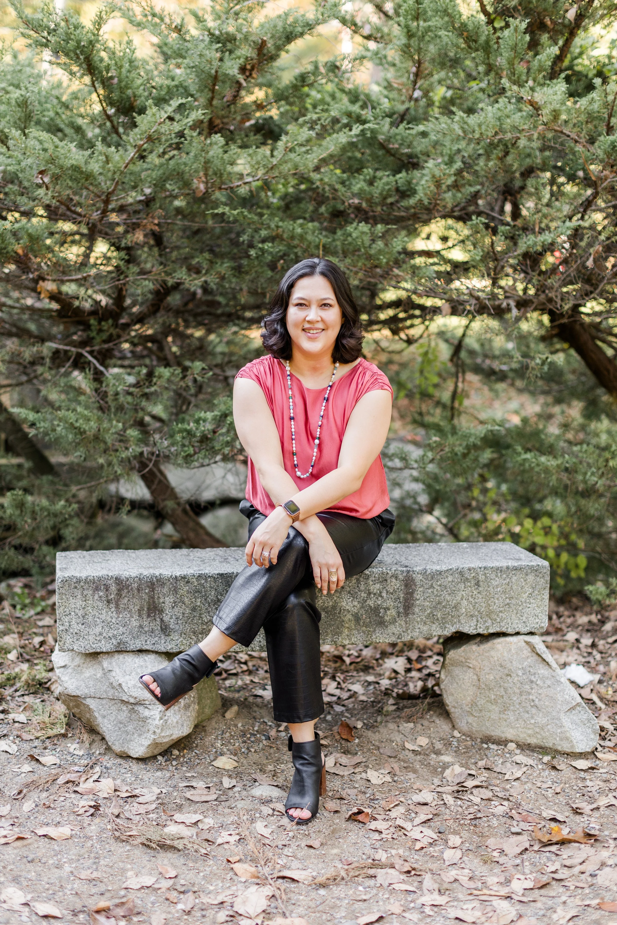 A woman with shoulder-length dark hair sitting on a stone bench outdoors, smiling, wearing a red sleeveless top, black leather pants, black open-toe boots, a colorful beaded necklace, and a smartwatch, with green trees and fallen leaves in the background.