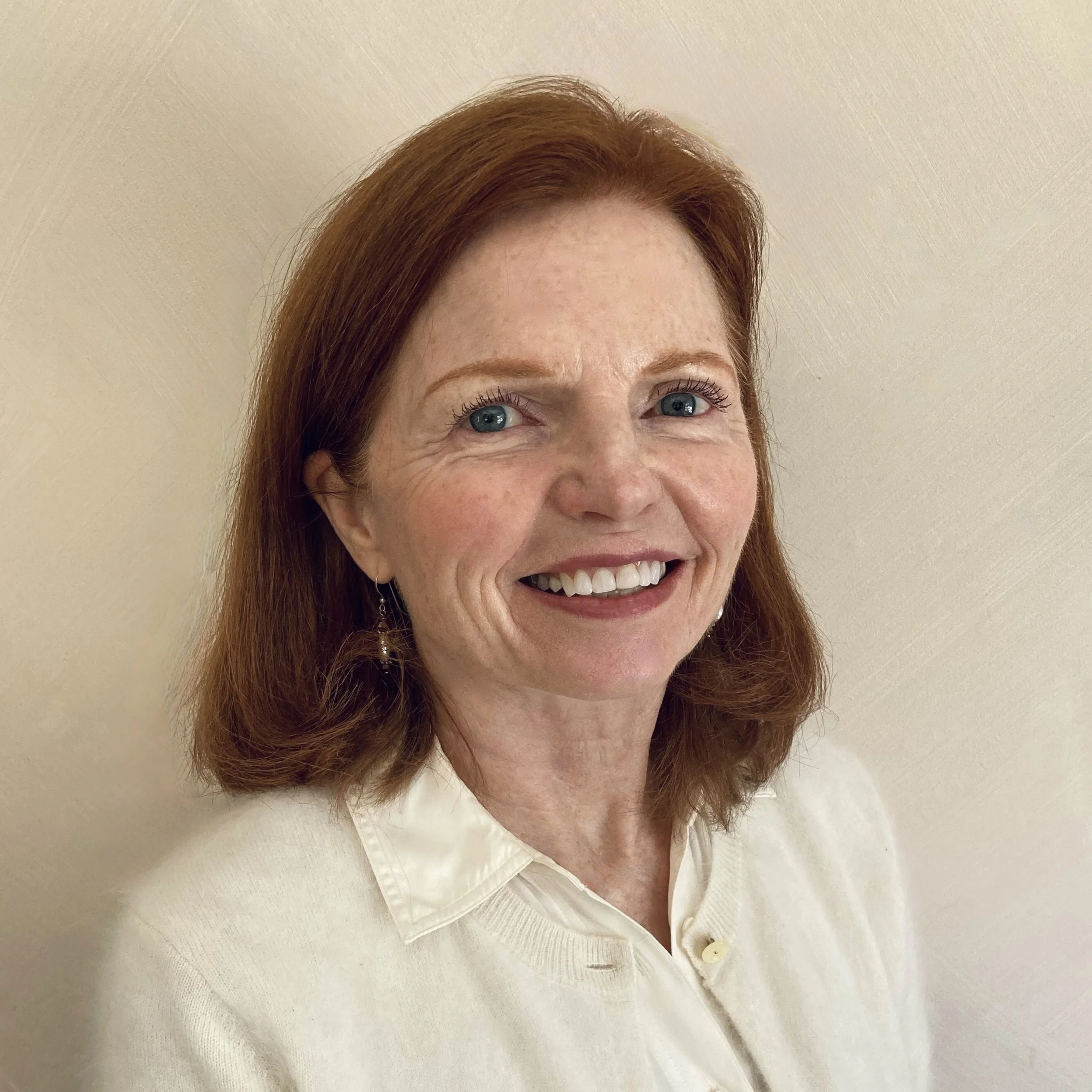 Close-up of a smiling woman with red hair wearing a white shirt, standing against a cream-colored wall.