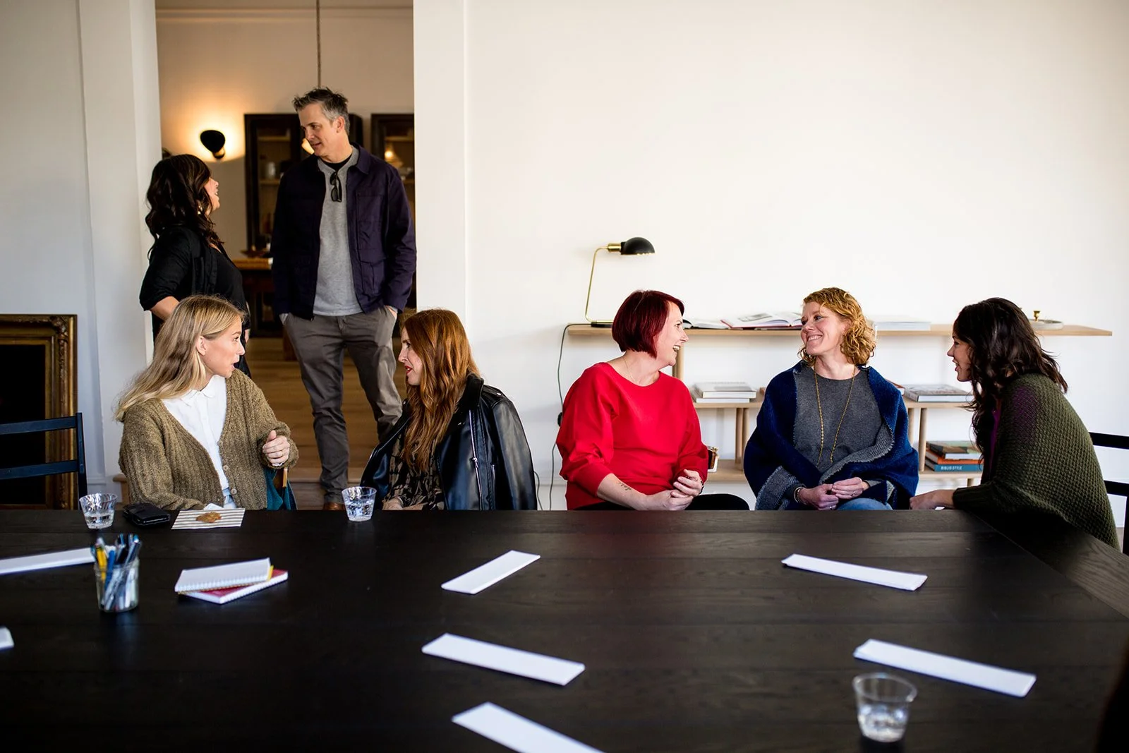 Six women and two men in a meeting room, some seated at a table and others standing, engaging in conversation.