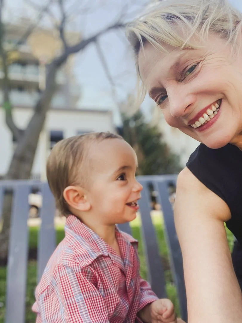 A woman and a young child smiling and looking at each other outdoors in a park or yard with trees and buildings in the background.