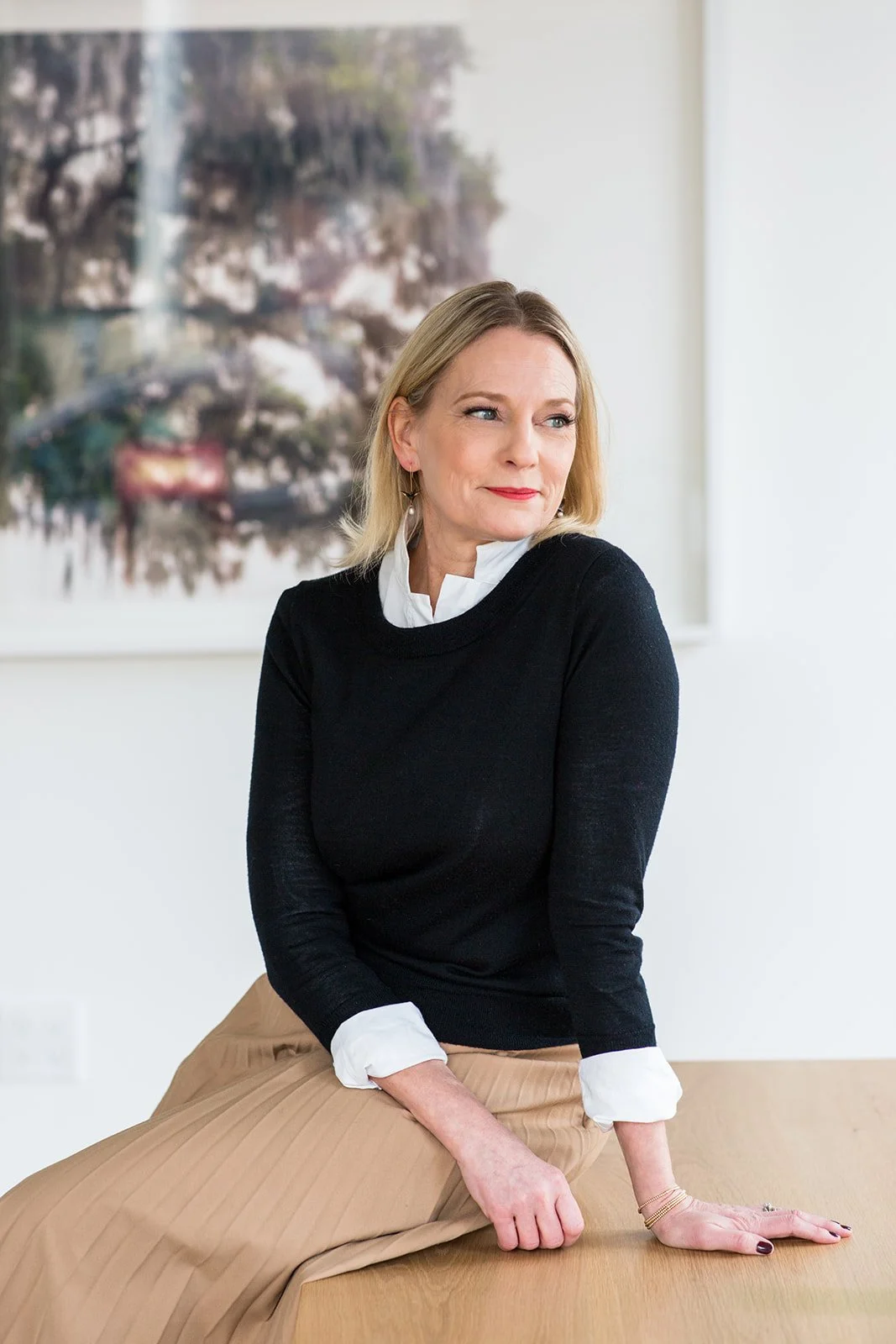 A woman with blonde hair sitting on a wooden table, wearing a black sweater with white cuffs and a white collar shirt underneath. She has a thoughtful expression and is looking to her left. There is a blurred painting or photograph of a waterfall and rocks in the background.