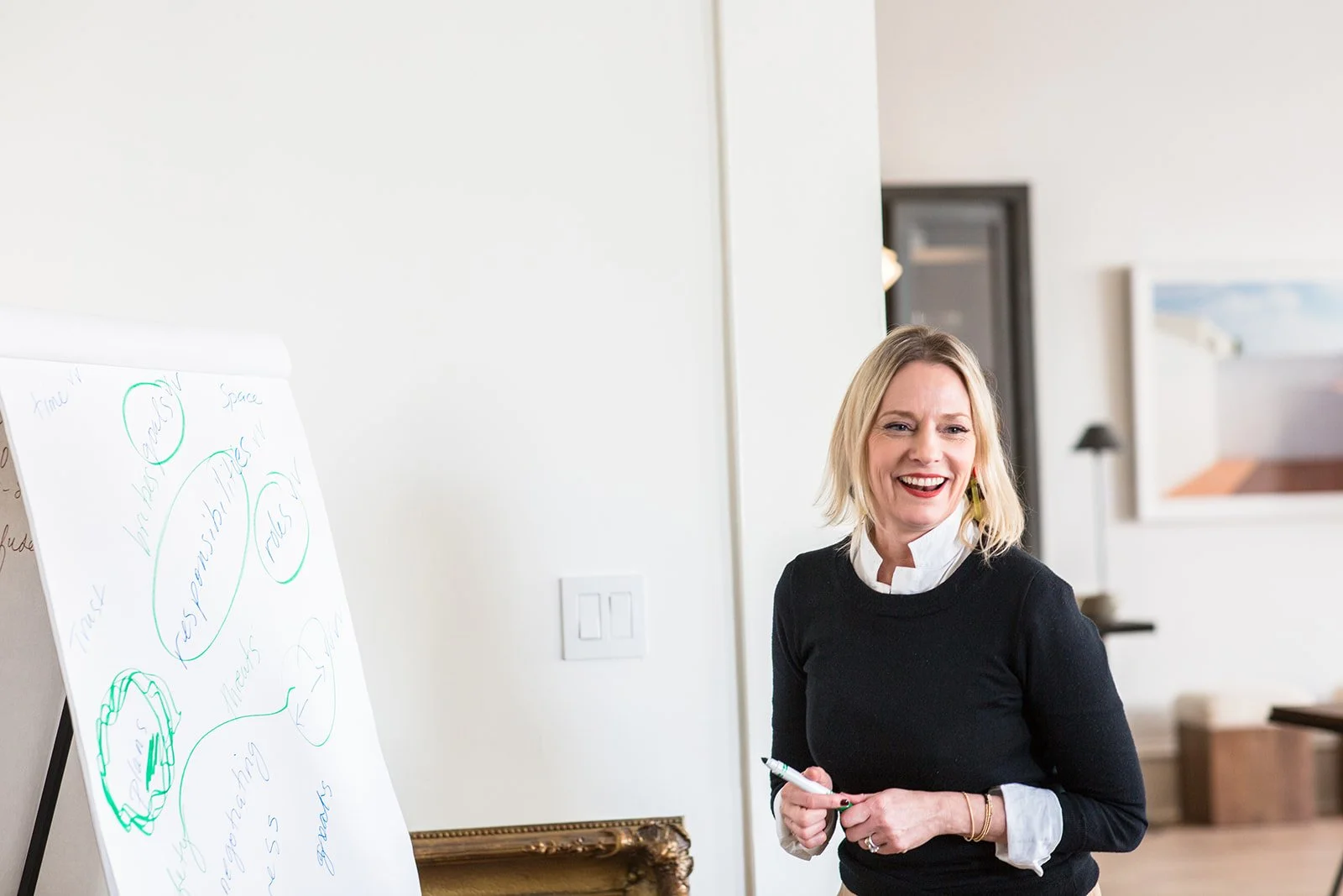 A woman smiling and holding a marker in a room with a whiteboard that has some writing and diagrams, and artwork on the walls.