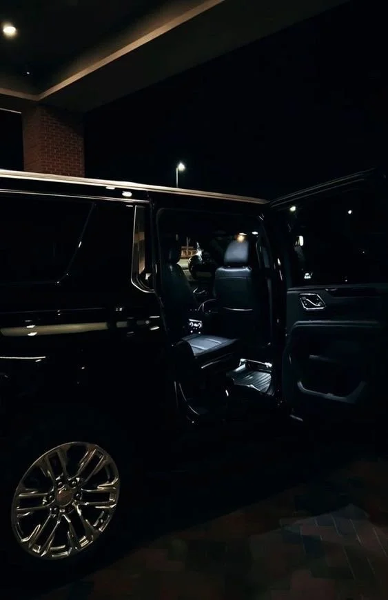 A black luxury SUV parked at night with the driver's side door open, showing the interior and part of the car's alloy wheel.
