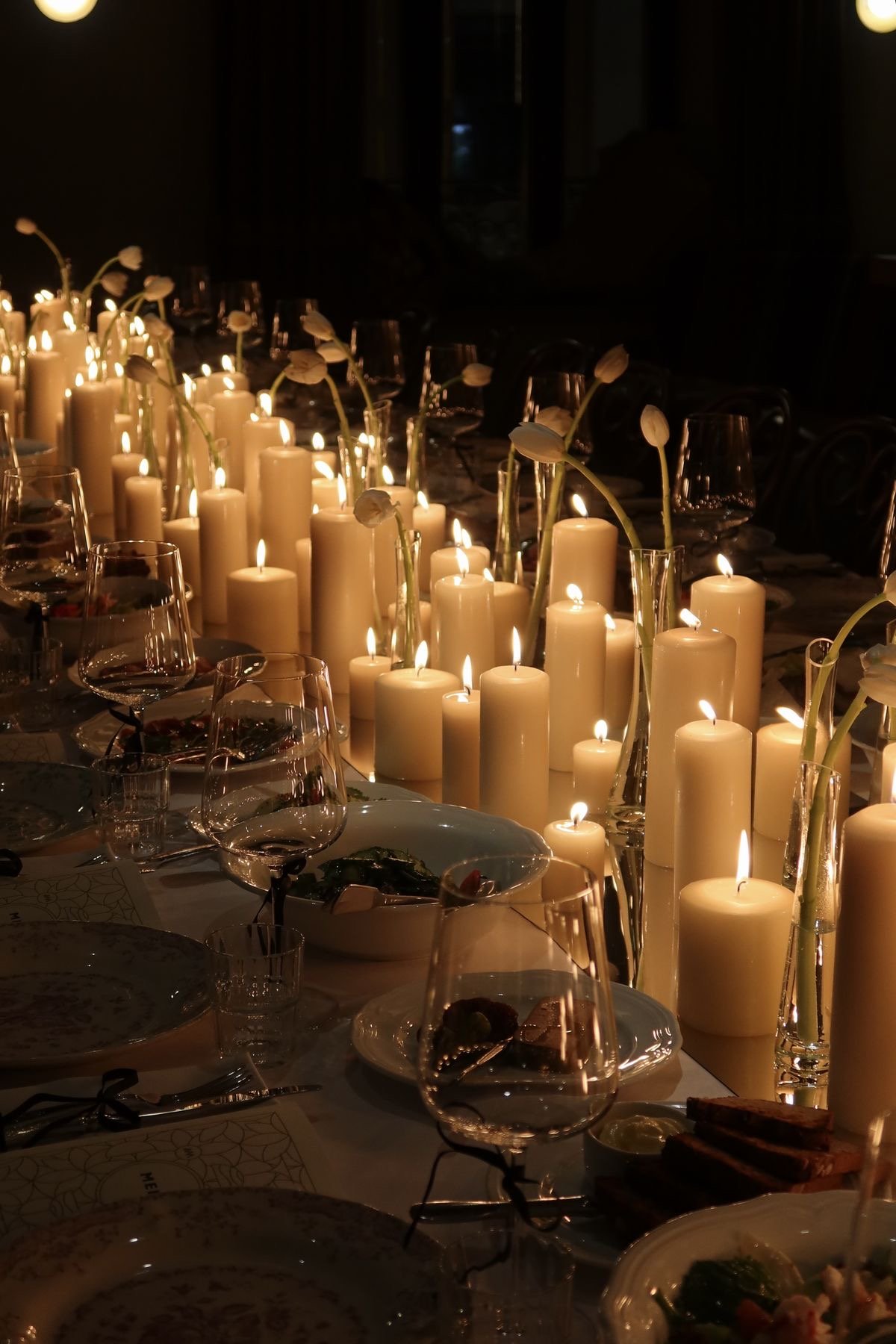 A long dinner table decorated with numerous lit candles, white flowers in vases, wine glasses, plates, and food.
